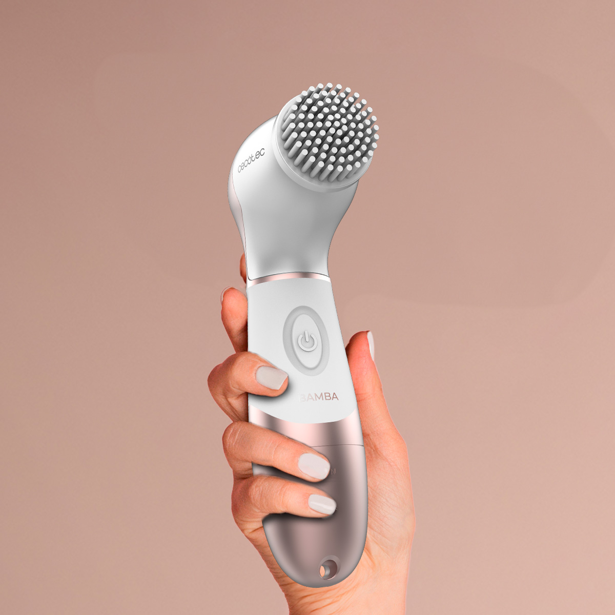 Una mano femenina sostiene un cepillo de limpieza facial blanco y oro rosa. El cabezal del cepillo tiene pequeñas cerdas. Fondo melocotón suave.