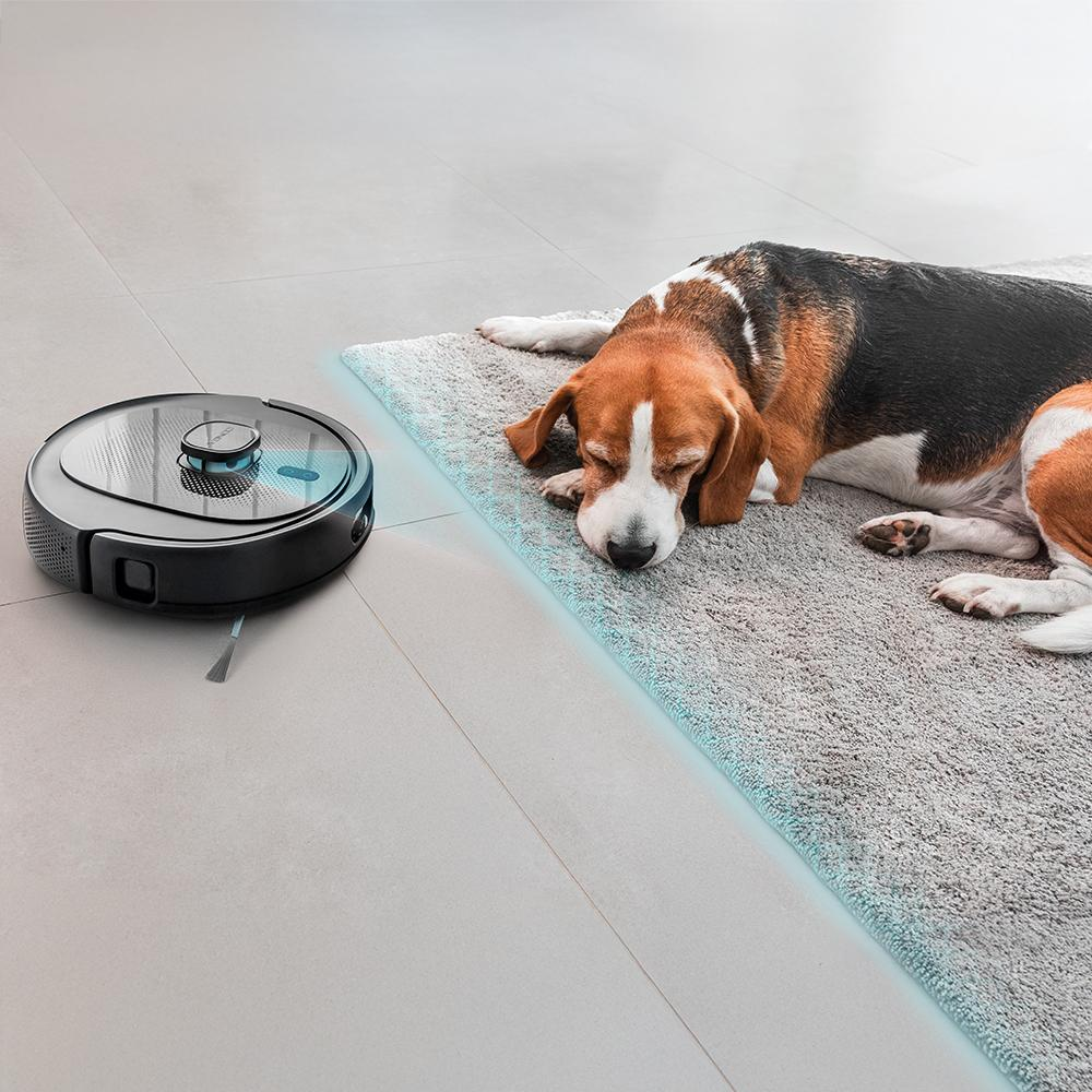 Un perro beagle duerme sobre una alfombra, junto a un robot aspirador en un suelo embaldosado. La alfombra tiene un borde azul claro.