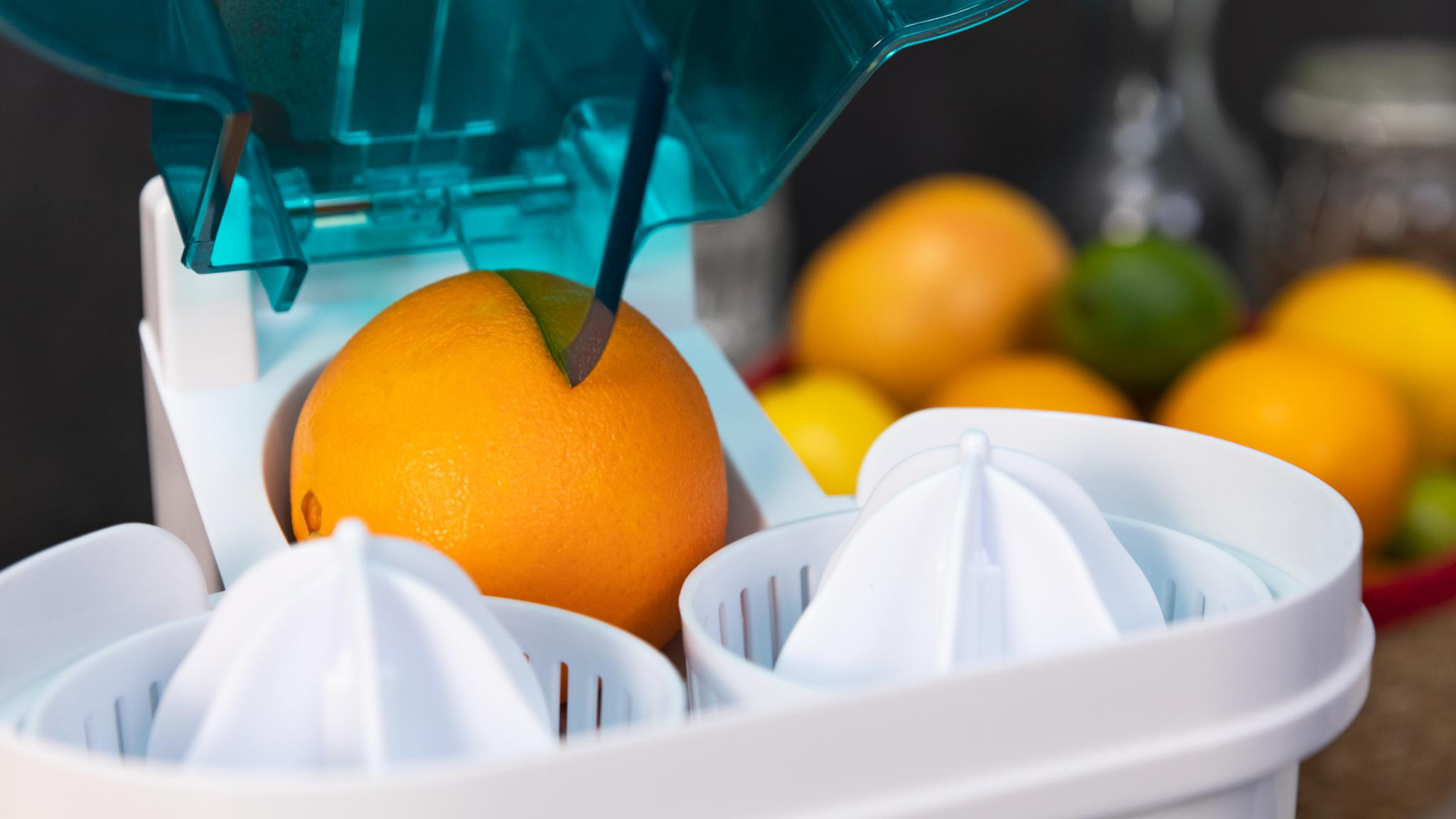 Una naranja se está exprimiendo en una exprimidora blanca. En el fondo se ven otras naranjas, limas y un limón.