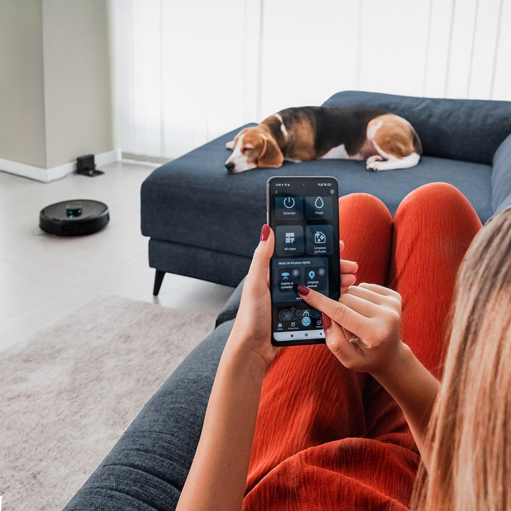 Mujer usando un teléfono para controlar una aspiradora robot. Un perro descansa en un sofá azul. Aspiradora en el fondo.