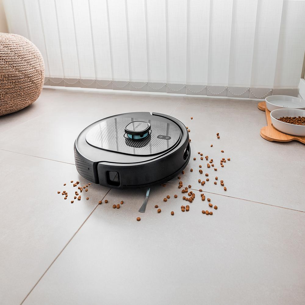 Una aspiradora robot negra limpia un suelo embaldosado salpicado de comida para mascotas. También hay un otomano tejido y cuencos para mascotas.