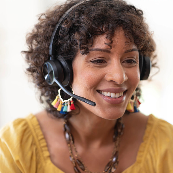Eine lächelnde Frau mit Headset, gelbem Oberteil und bunten Ohrringen. Sie scheint sich in einem Videoanruf oder in einer Kundendienstrolle zu befinden.