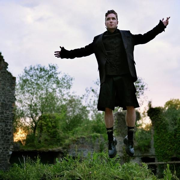 Mann in schwarzem Anzug und Kilt schwebt über Gras, Arme ausgestreckt. Ruinen und Bäume im Hintergrund, Himmel darüber.