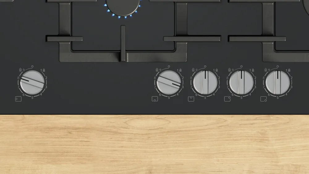 Zbliżenie czarnej płyty grzewczej ze srebrnymi pokrętłami. Płyta grzewcza znajduje się na jasnej drewnianej powierzchni. Jeden palnik jest zapalony niebieskimi płomieniami.
