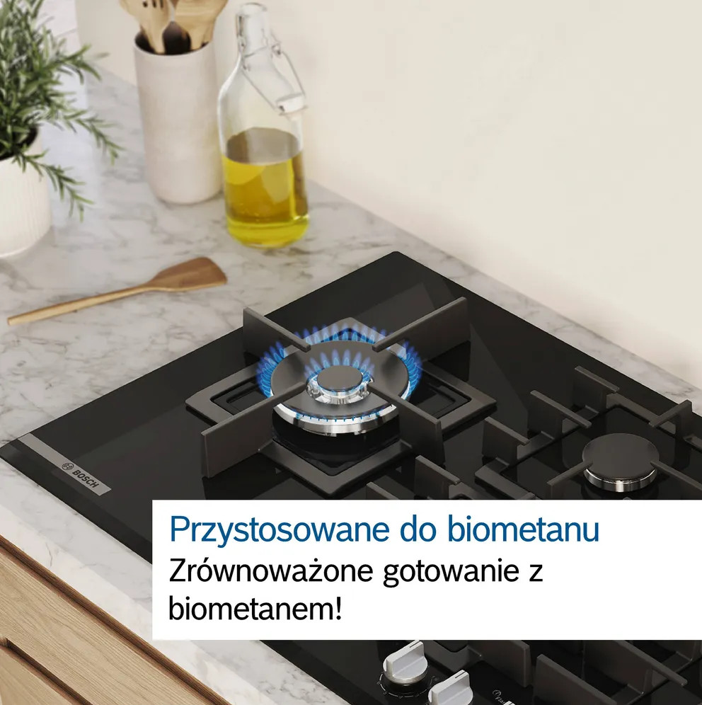 Czarna płyta gazowa z niebieskim płomieniem jest zapalona. Szklana butelka z olejem, drewniana łyżka i rośliny w tle.