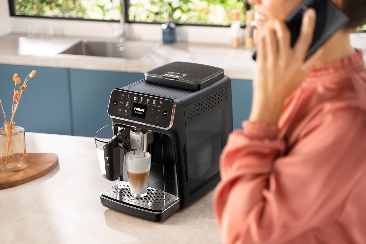 Una cafetera espresso Philips negra está preparando café. Una persona está hablando por teléfono. Un vaso de café está en la bandeja.