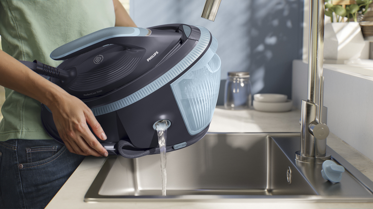 Una persona vierte agua de una plancha Philips en un fregadero. La plancha es negra y azul. La escena es brillante.