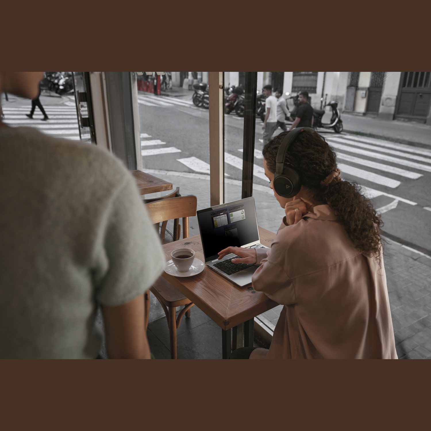 Vrouw in café met koptelefoon gebruikt laptop. Koffie op tafel. Straatbeeld zichtbaar door het raam. Een andere persoon op de voorgrond.
