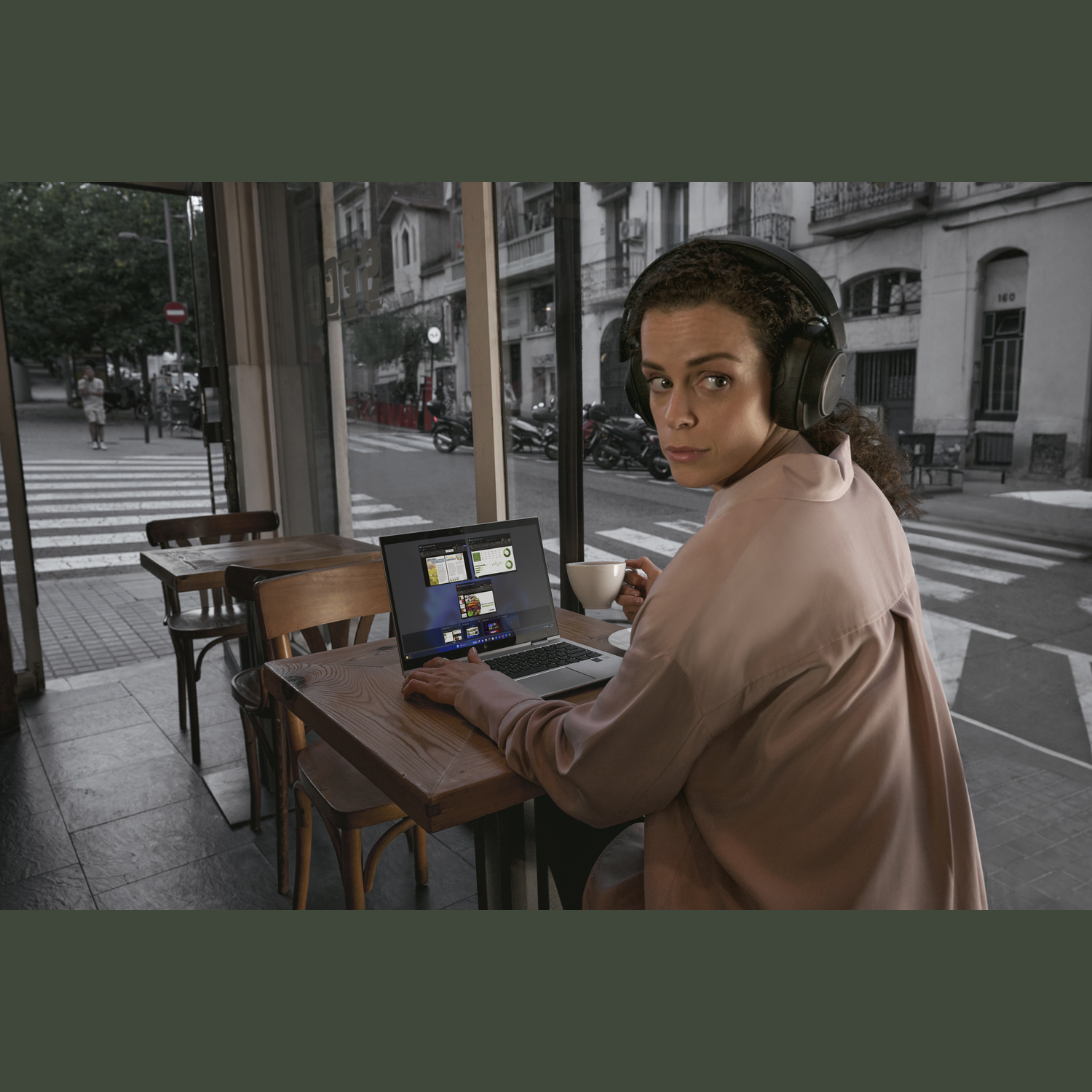 Vrouw met koptelefoon, gebruikt een laptop aan een cafétafel, kijkt over haar schouder. Straattafereel zichtbaar door het raam.