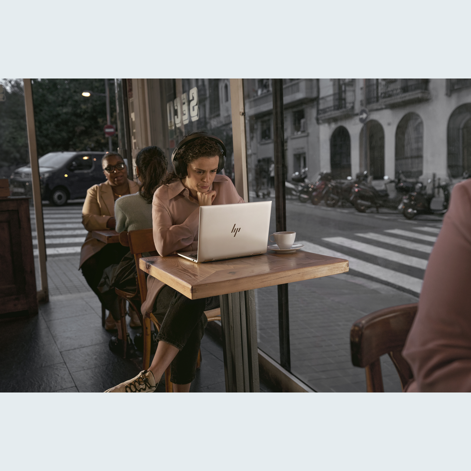 Woman in cafe uses laptop, wearing headphones. Two others sit nearby. Outside, a street with cars and parked scooters is visible.