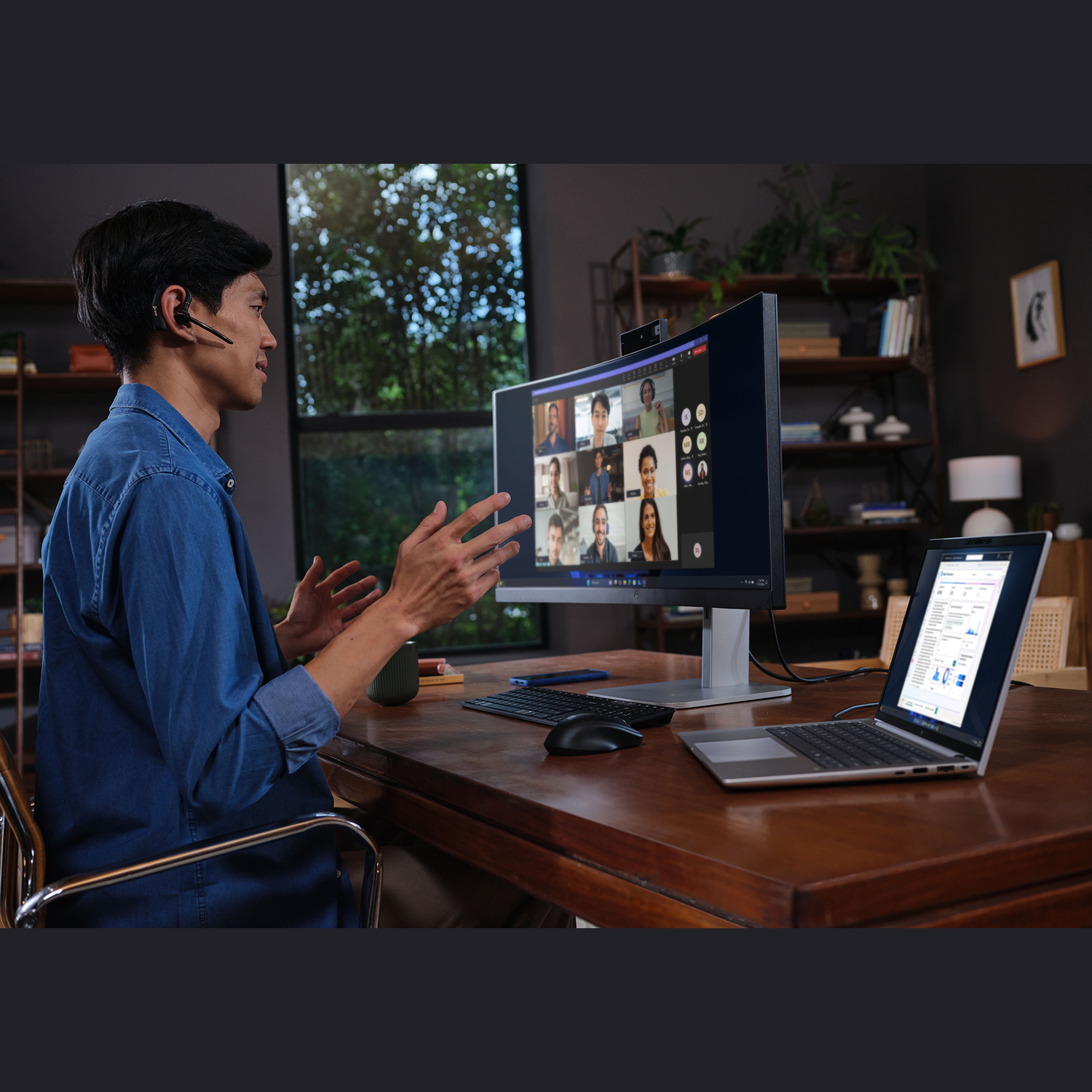 Een man met een headset gebaart tijdens een videogesprek. Een grote monitor en een laptop tonen de deelnemers. Houten bureau en thuiskantoor.