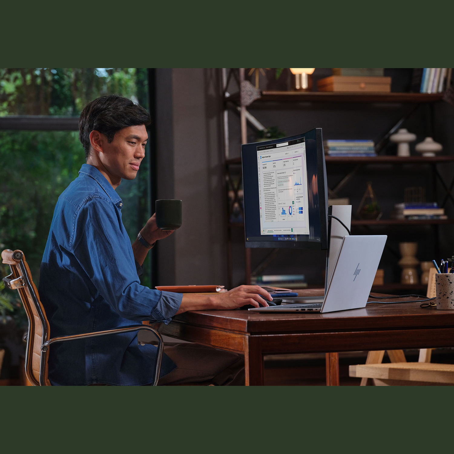 Een man in een blauw overhemd zit aan een bureau, houdt een mok vast en werkt op een laptop en monitor. Een boekenplank op de achtergrond.