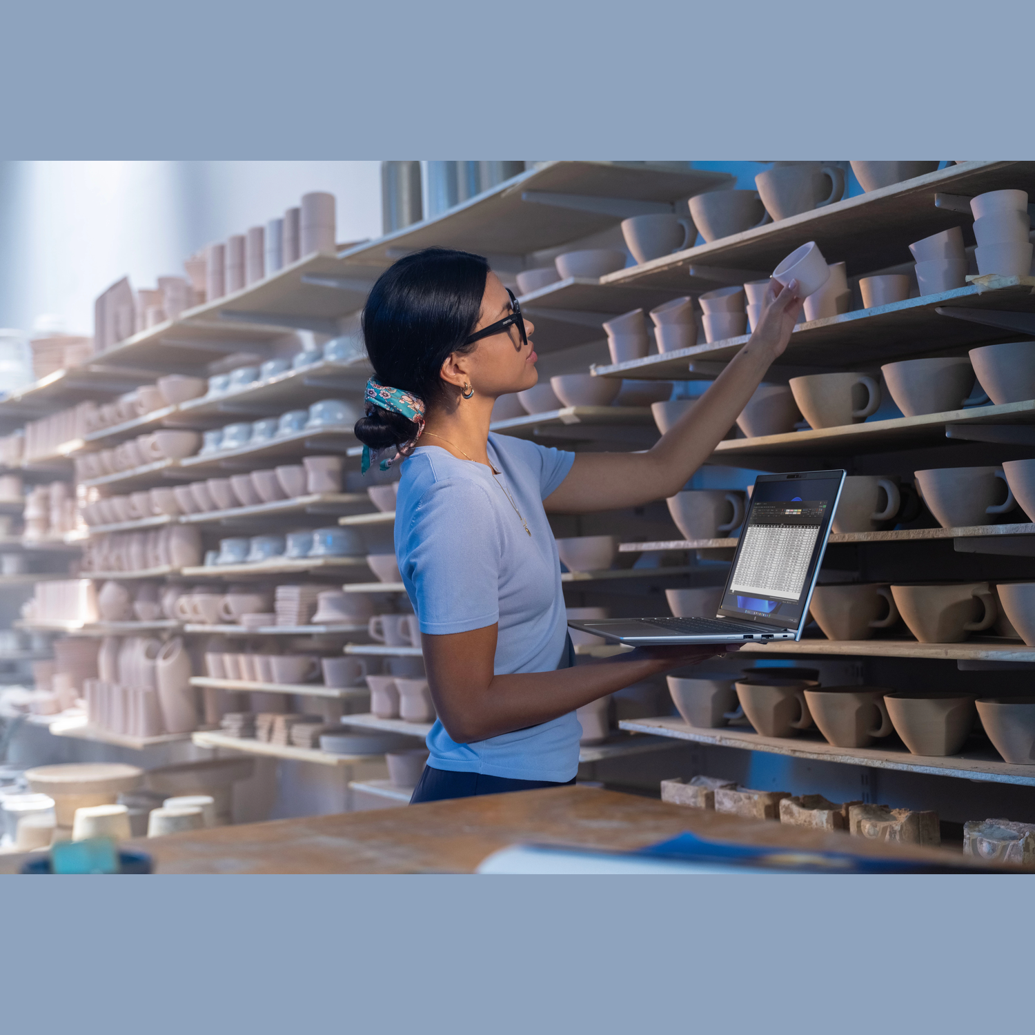 Woman in glasses examines pottery on shelves, holding a laptop. Cups and bowls are on the shelves. Pottery studio setting.