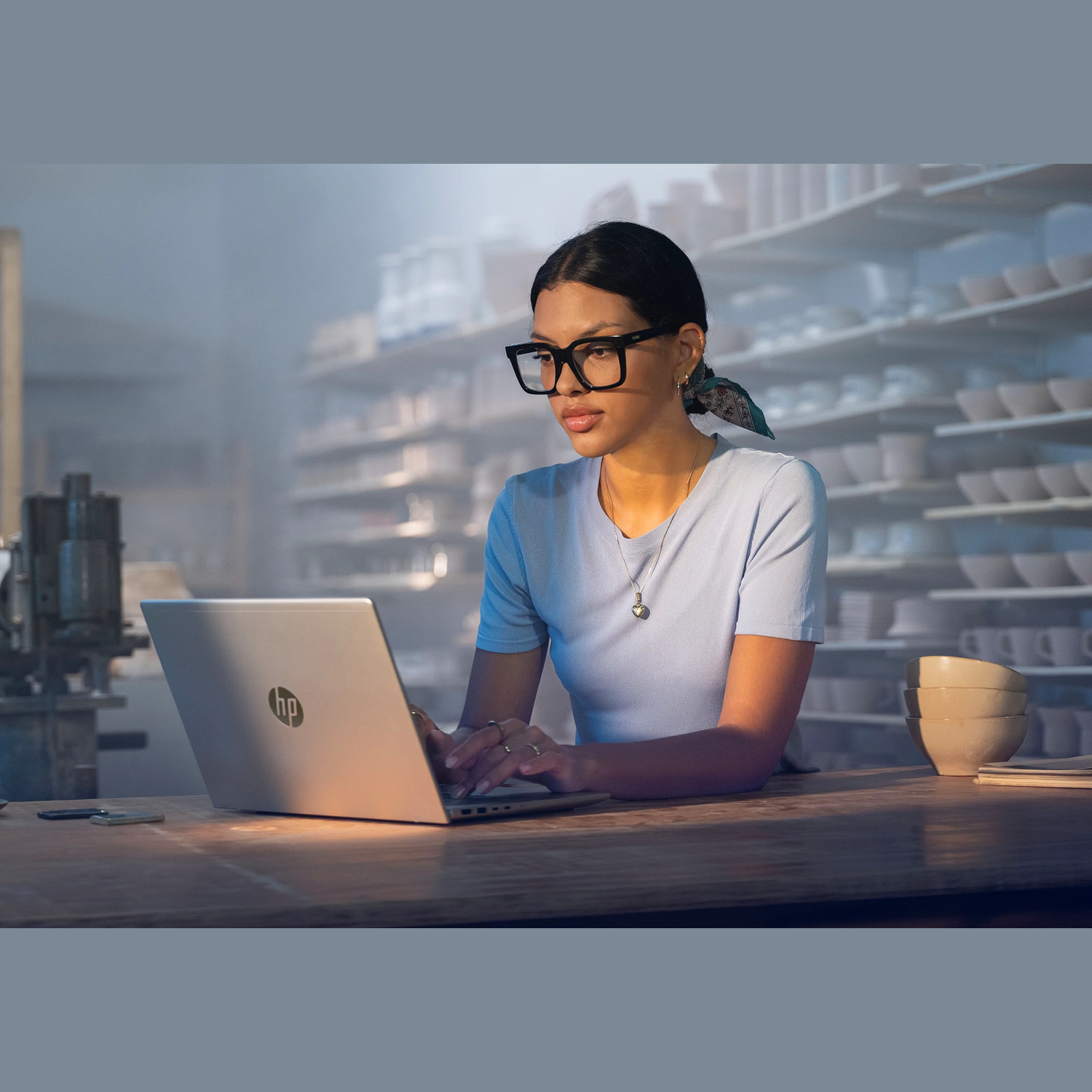 A woman with glasses types on an HP laptop. She is in a pottery studio. Background shows shelves of pottery.