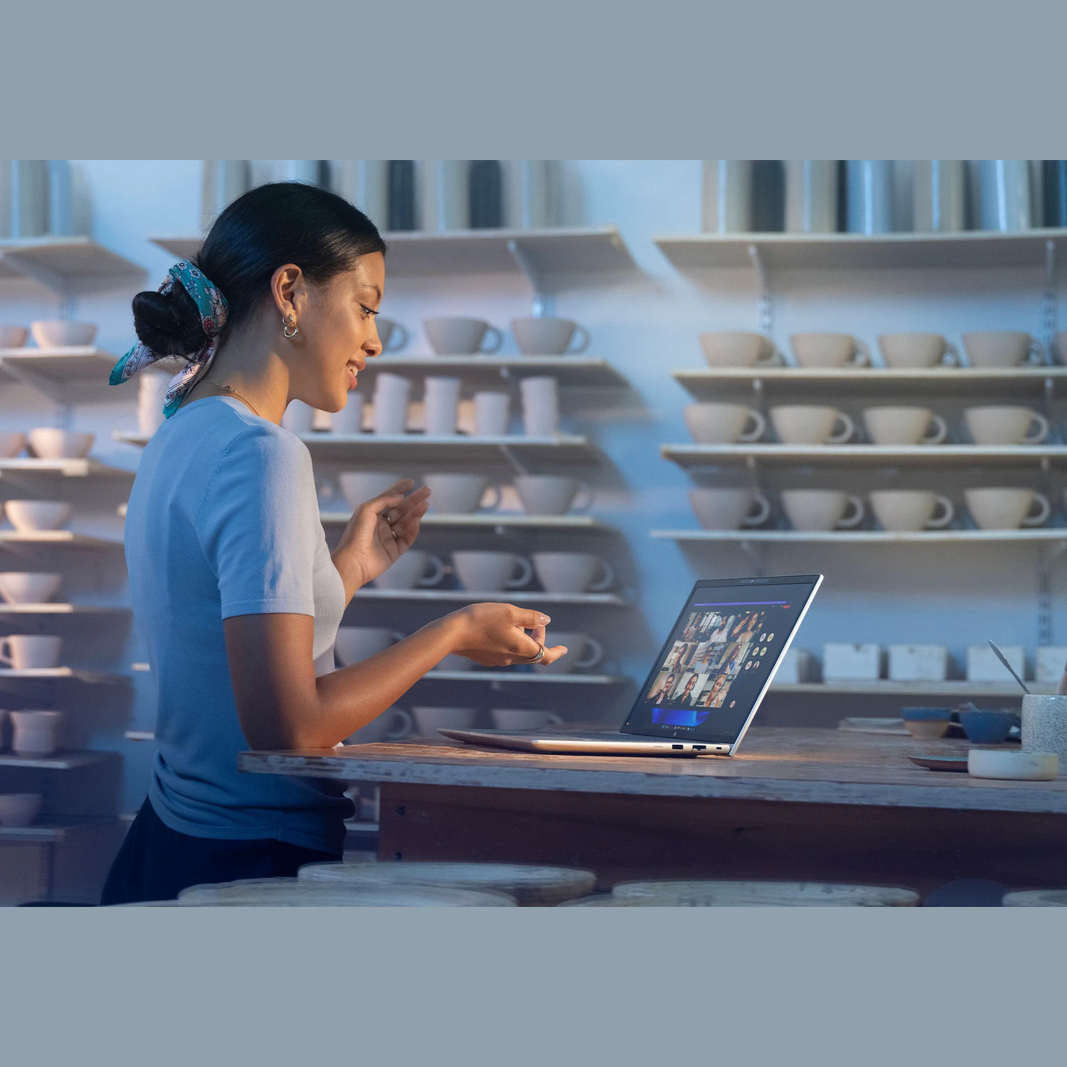 Woman in pottery studio video calls on laptop. Shelves hold pottery. She gestures towards screen.
