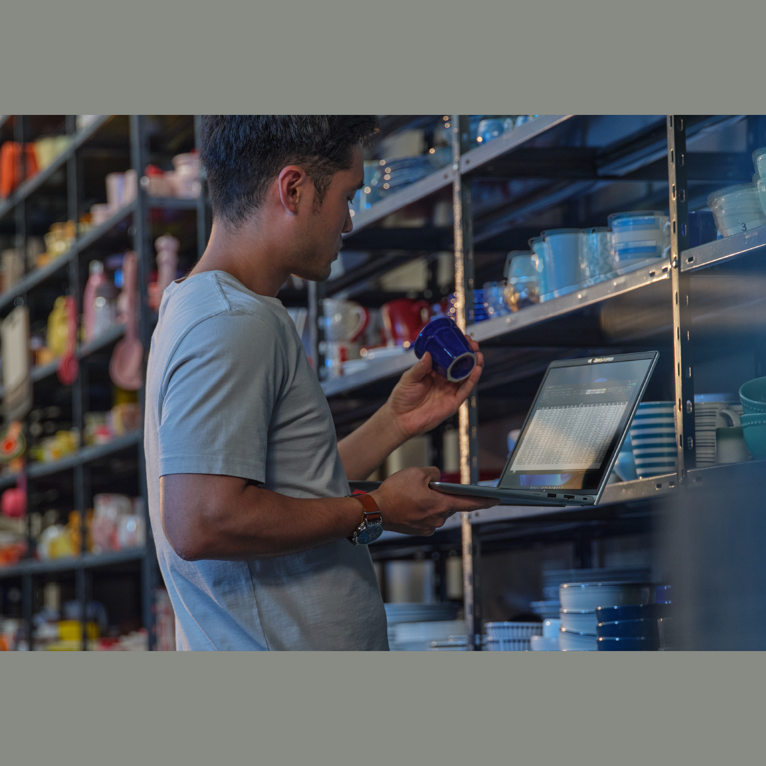 Man in a store examines a blue cup while using a laptop. Shelves are filled with dishes of various colors and styles.