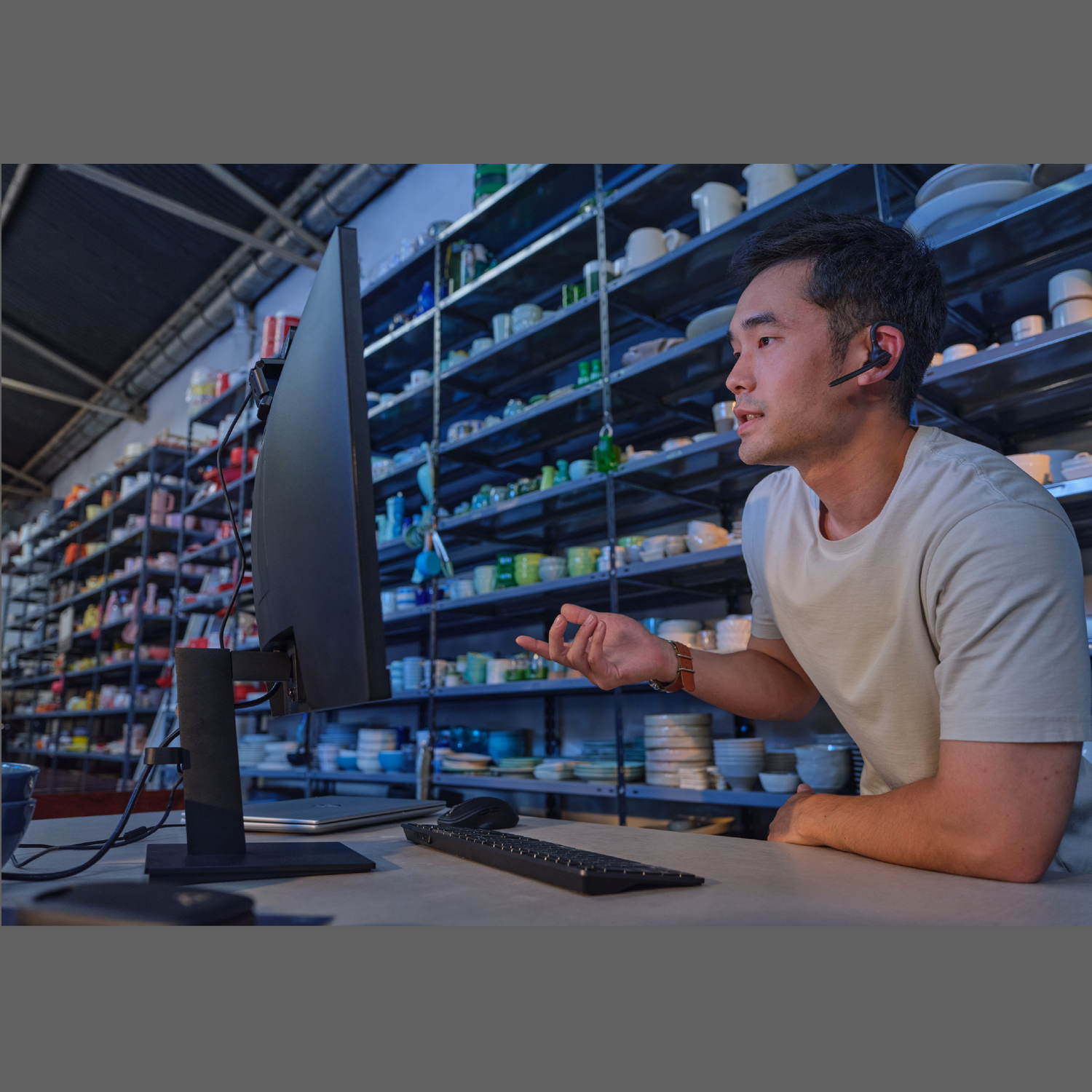 A man with a headset gestures while looking at a computer screen. Shelves filled with dishware are in the background.