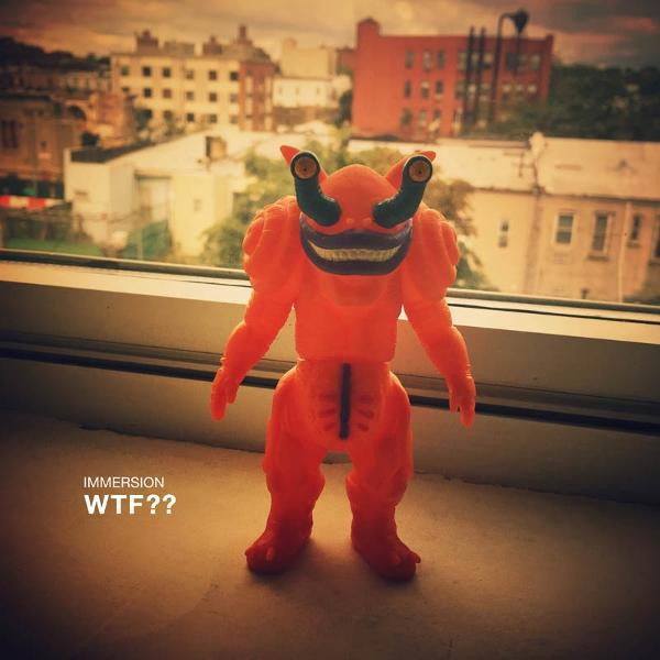 Ein orangefarbenes Monstertoy mit großen Augen und einem grinsenden Mund steht auf einer Fensterbank, im Hintergrund sind Stadtgebäude zu sehen.
