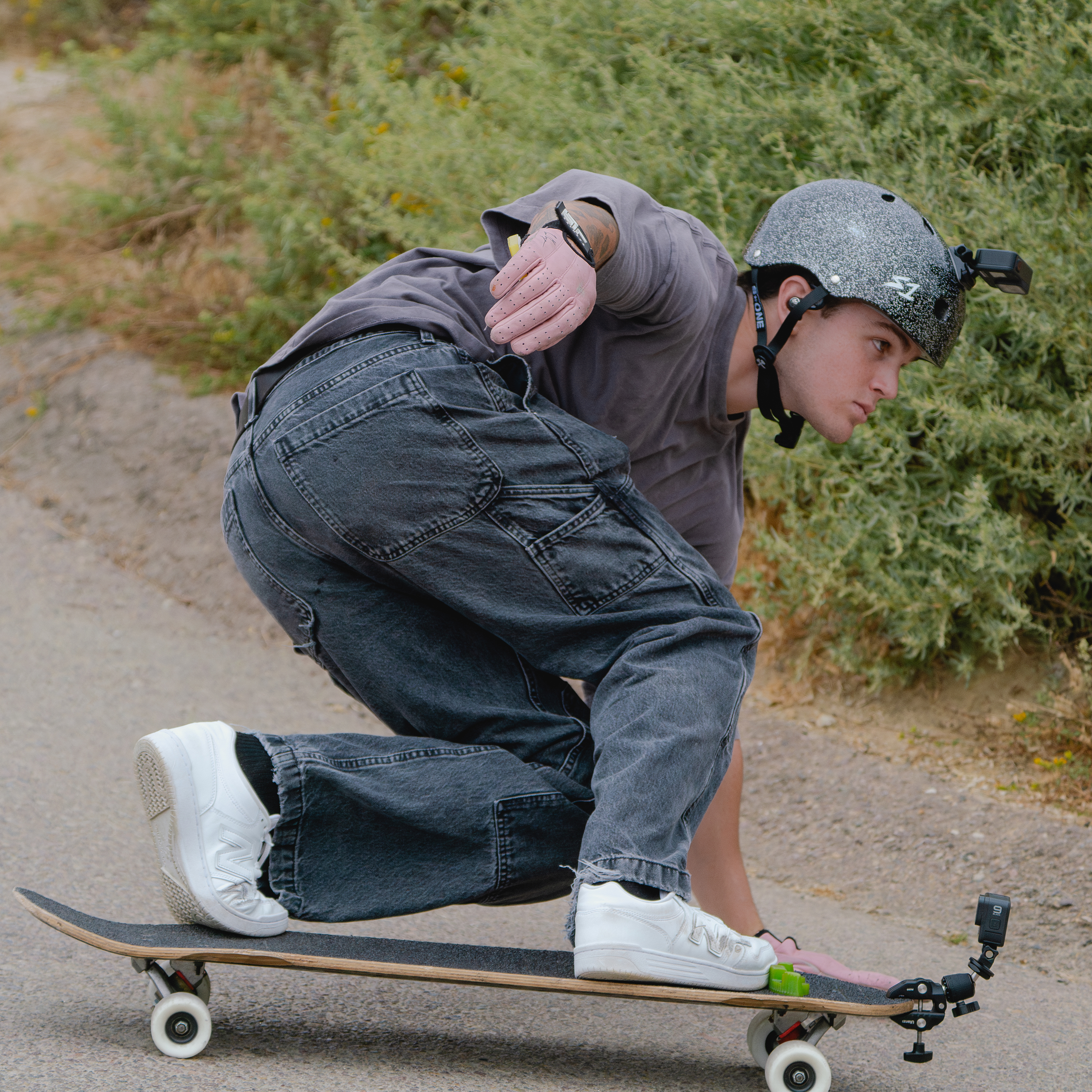 Eine Person auf einem Skateboard lehnt sich nach vorne und trägt einen Helm. Grüne Büsche sind im Hintergrund.