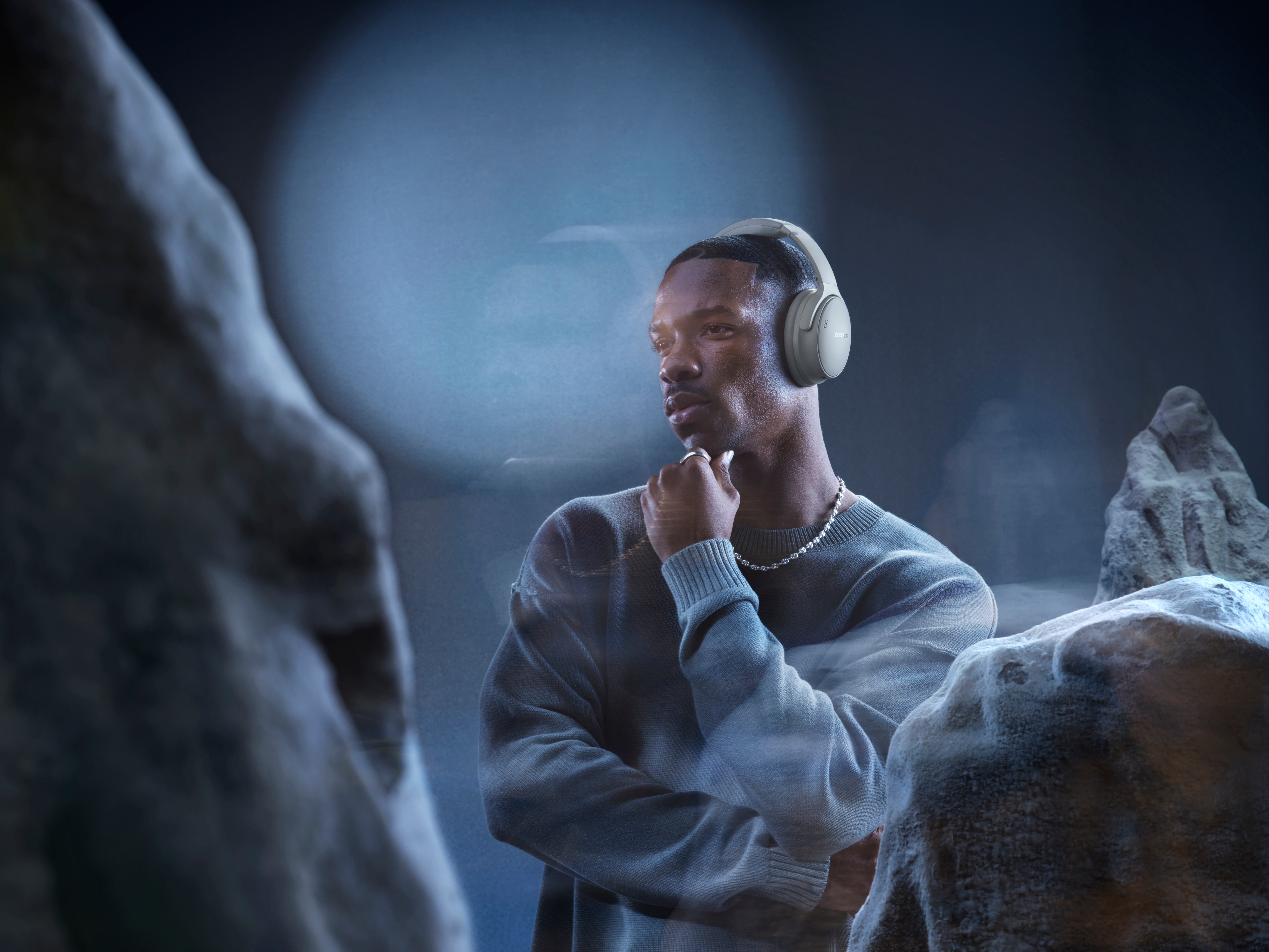 Un homme avec un casque, un pull et un collier, l'air pensif. Fond: formations rocheuses et lumière bleue.