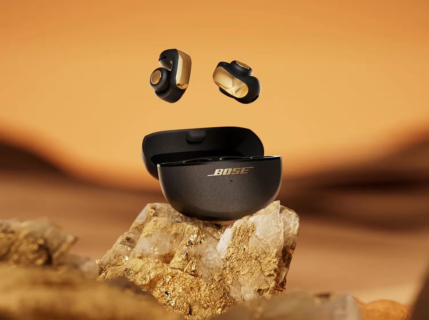 Auricolari Bose neri e oro fluttuanti sopra la custodia di ricarica aperta su una roccia. Lo sfondo è arancione.
