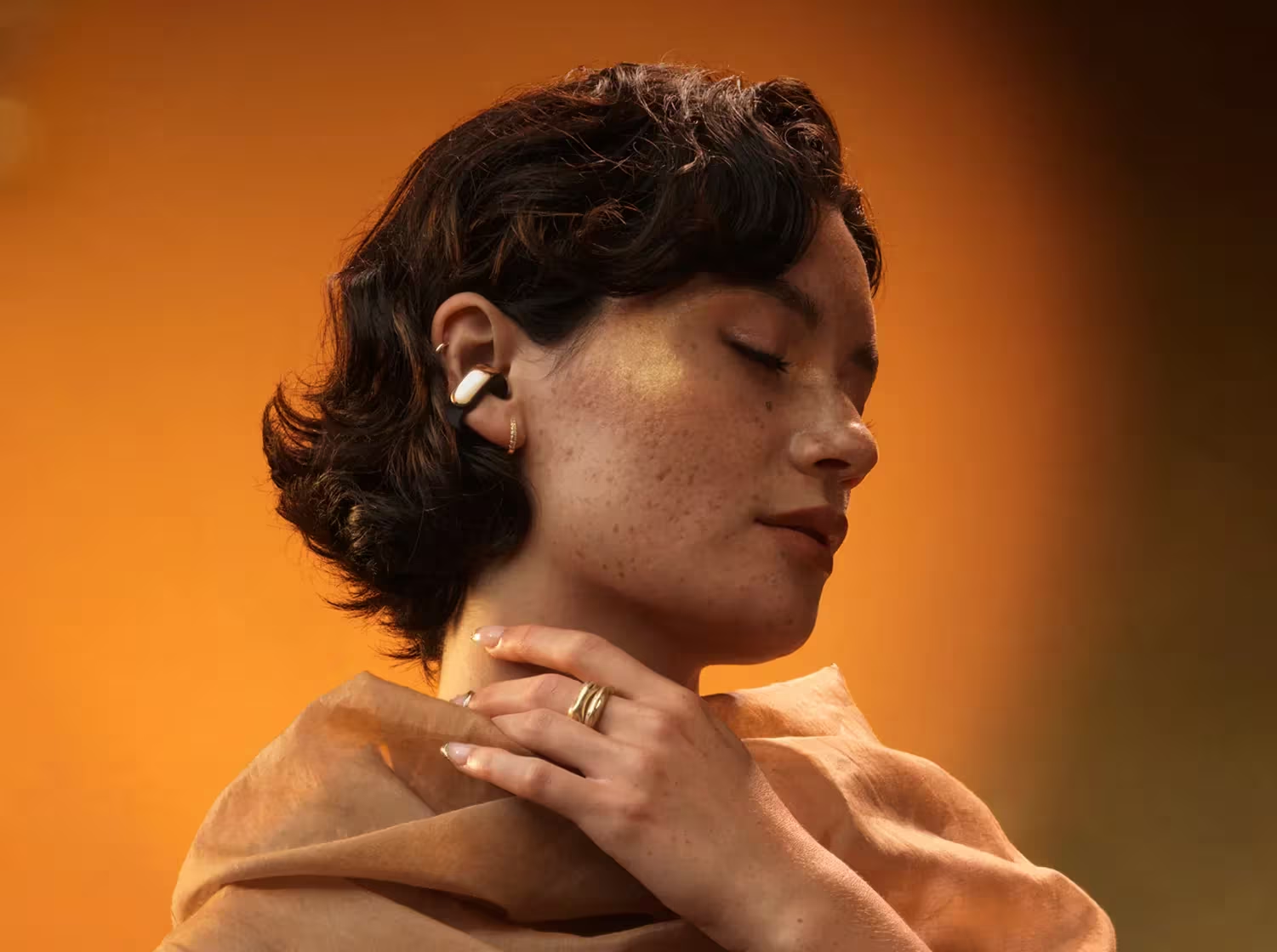 Una donna con lentiggini e occhi chiusi indossa un auricolare. Indossa anelli e uno scialle, con uno sfondo arancione caldo.