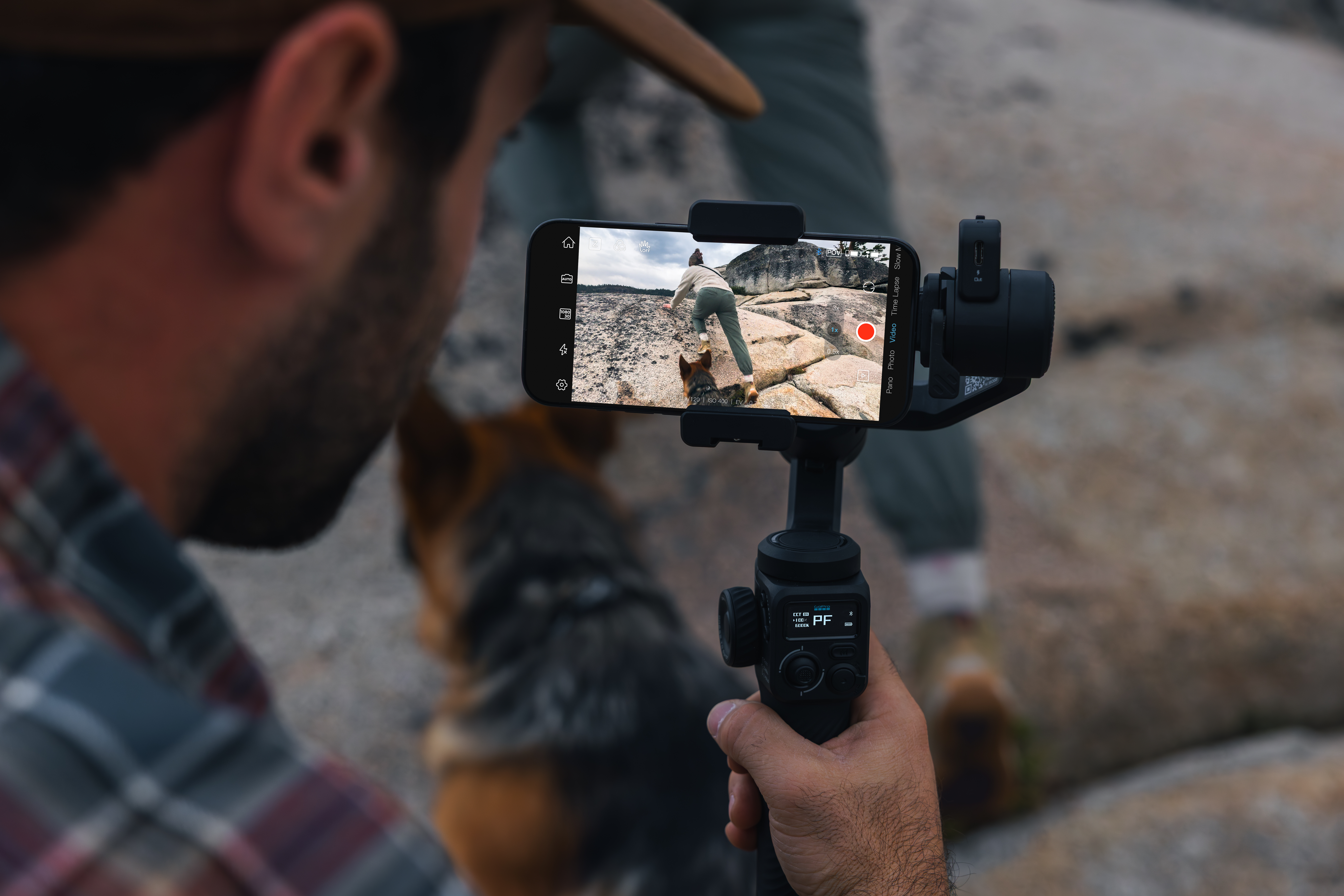 Eine Person nimmt ein Video mit einem an einem Gimbal befestigten Telefon auf, mit einem Hund im Vordergrund und einer Felslandschaft auf dem Bildschirm.
