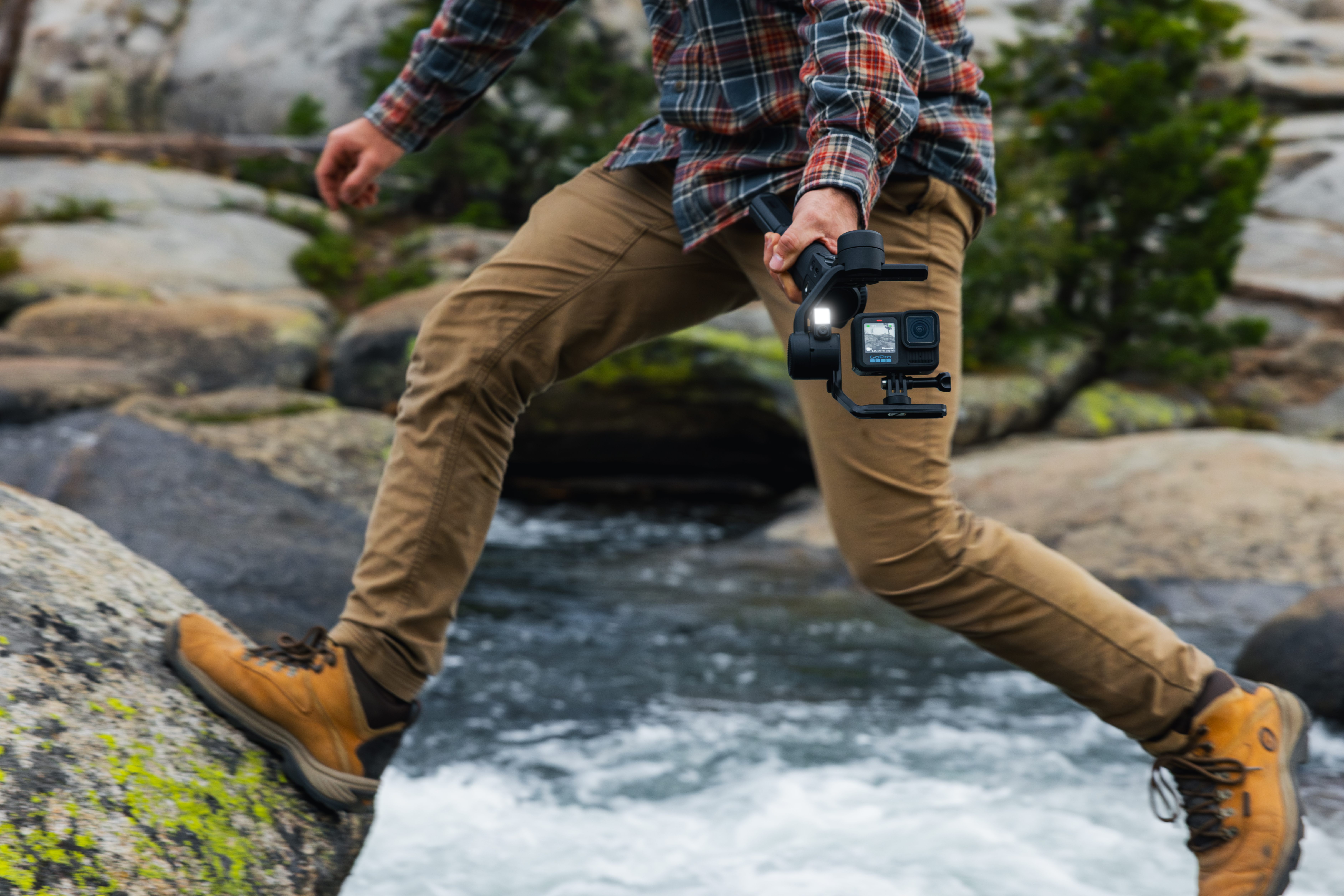 Person springt über Felsen, hält Kamera, trägt kariertes Hemd und braune Hose mit Stiefeln. Hintergrund: Wasser und Bäume.