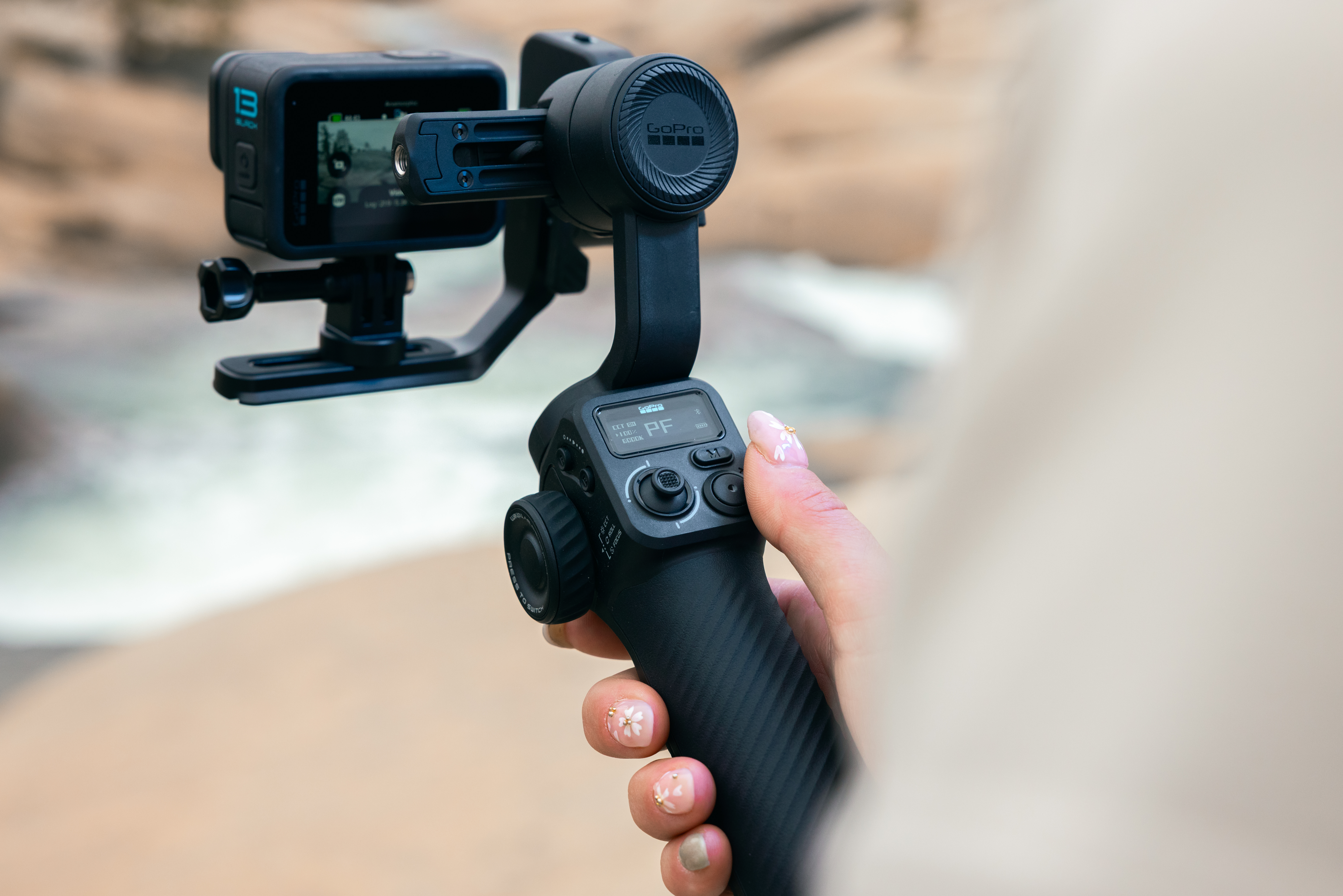 Eine Person hält eine schwarze GoPro-Kamera an einem Stabilisator. Der Hintergrund ist unscharf mit einem Fluss und Felsen.