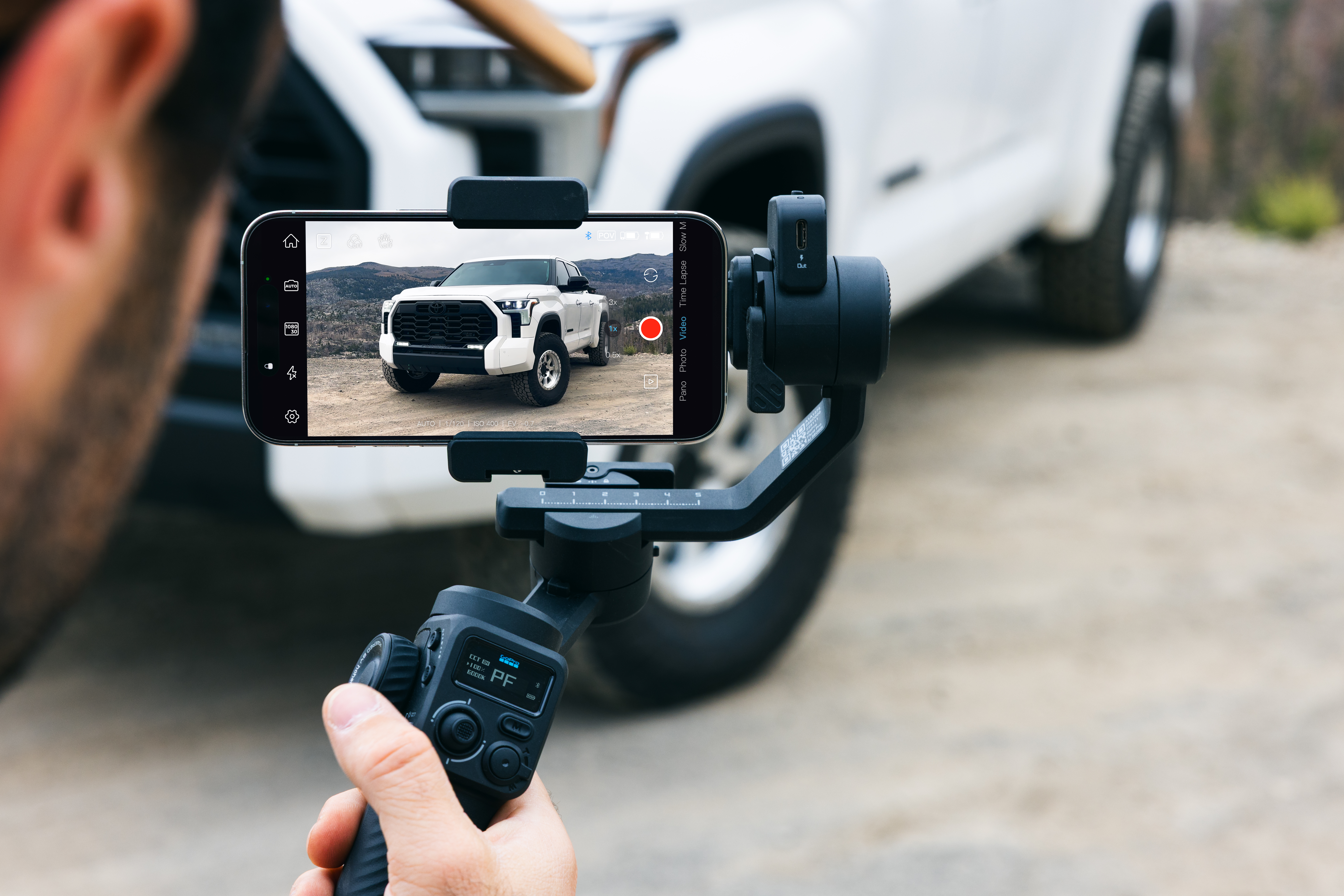 Eine Person filmt einen weißen LKW mit einem Telefon, das auf einem Gimbal montiert ist. Der Hintergrund zeigt eine verschwommene Umgebung.