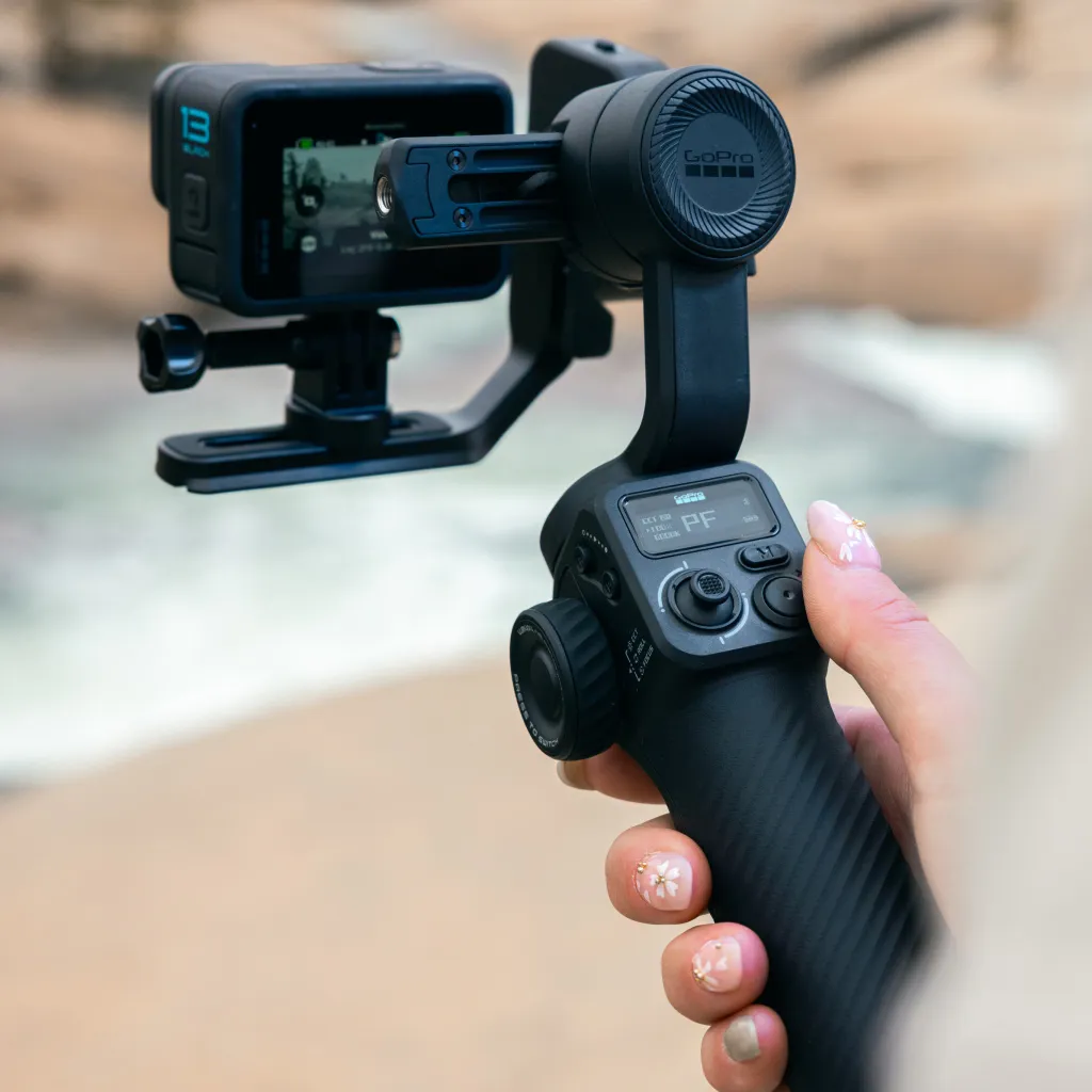 Egy személy egy fekete GoPro kamerát tart egy gimbalon. A képernyőn egy kép látható. A háttér elmosódott.