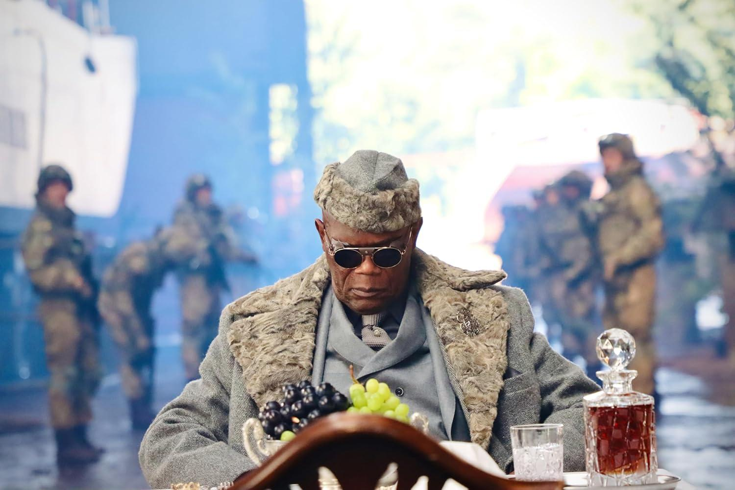 Mann mit Sonnenbrille, Pelzmantel und Hut. Trauben, eine Karaffe und Soldaten im Hintergrund.