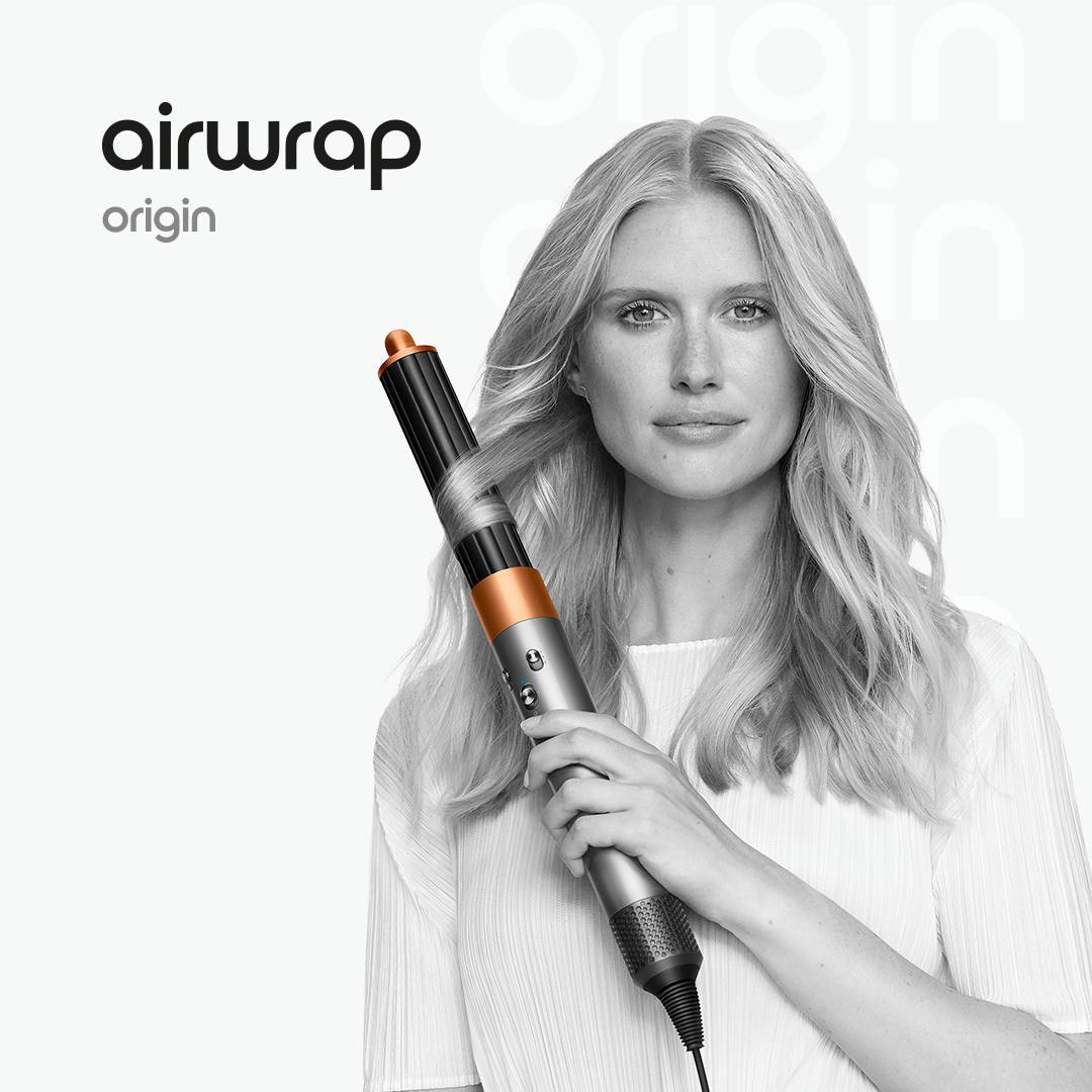 Mujer rubia sosteniendo una herramienta para peinar el cabello. La herramienta es negra y cobriza, y se está rizado el pelo. El fondo es blanco.