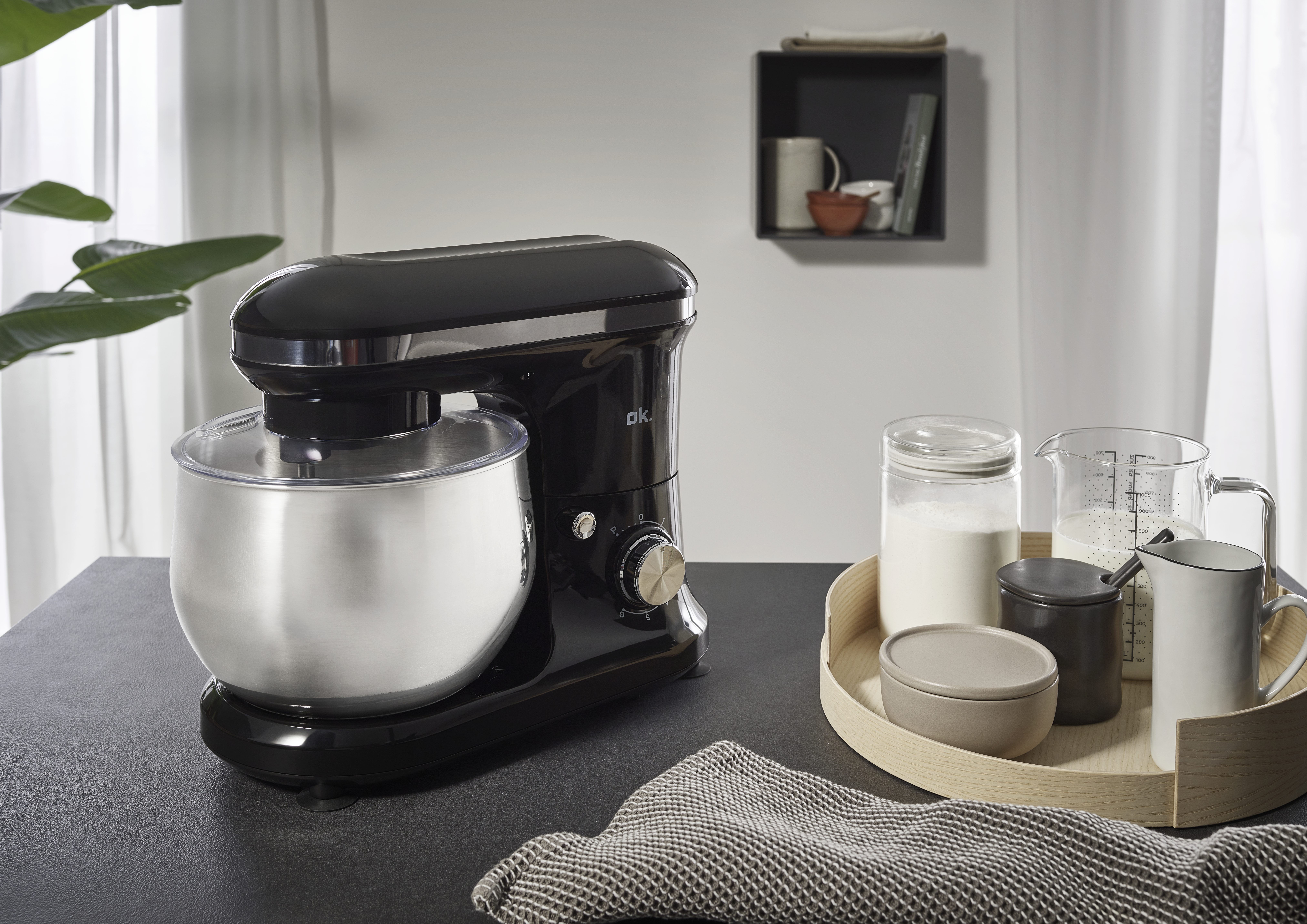 Ein schwarzer Küchenmixer steht auf einer dunklen Oberfläche mit Zutaten und einem Tablett. Eine grüne Pflanze und ein Regal sind im Hintergrund.