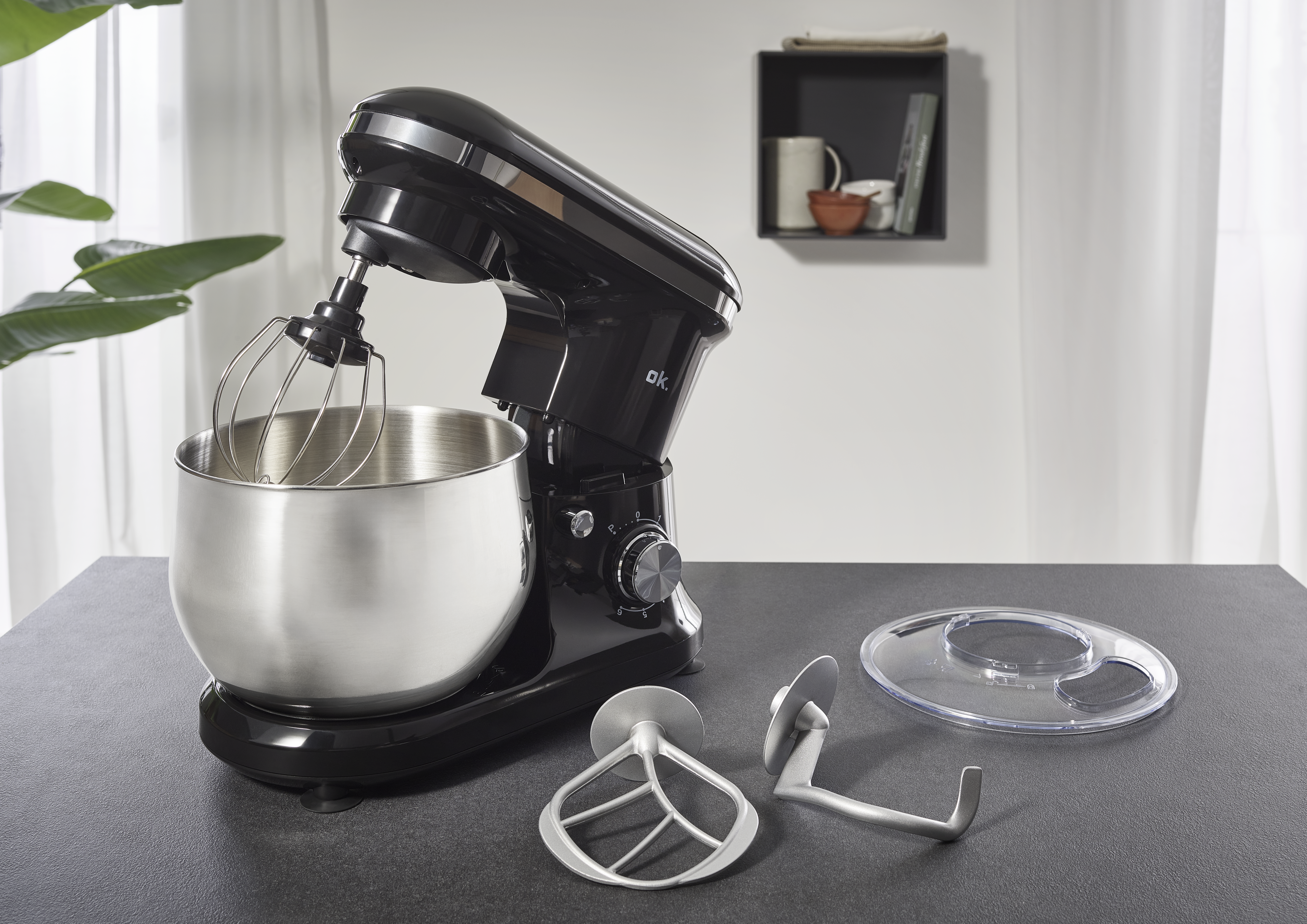 Ein schwarzer Küchenmixer mit Edelstahlschüssel und Aufsätzen auf einer schwarzen Oberfläche, im Hintergrund ein Fenster.