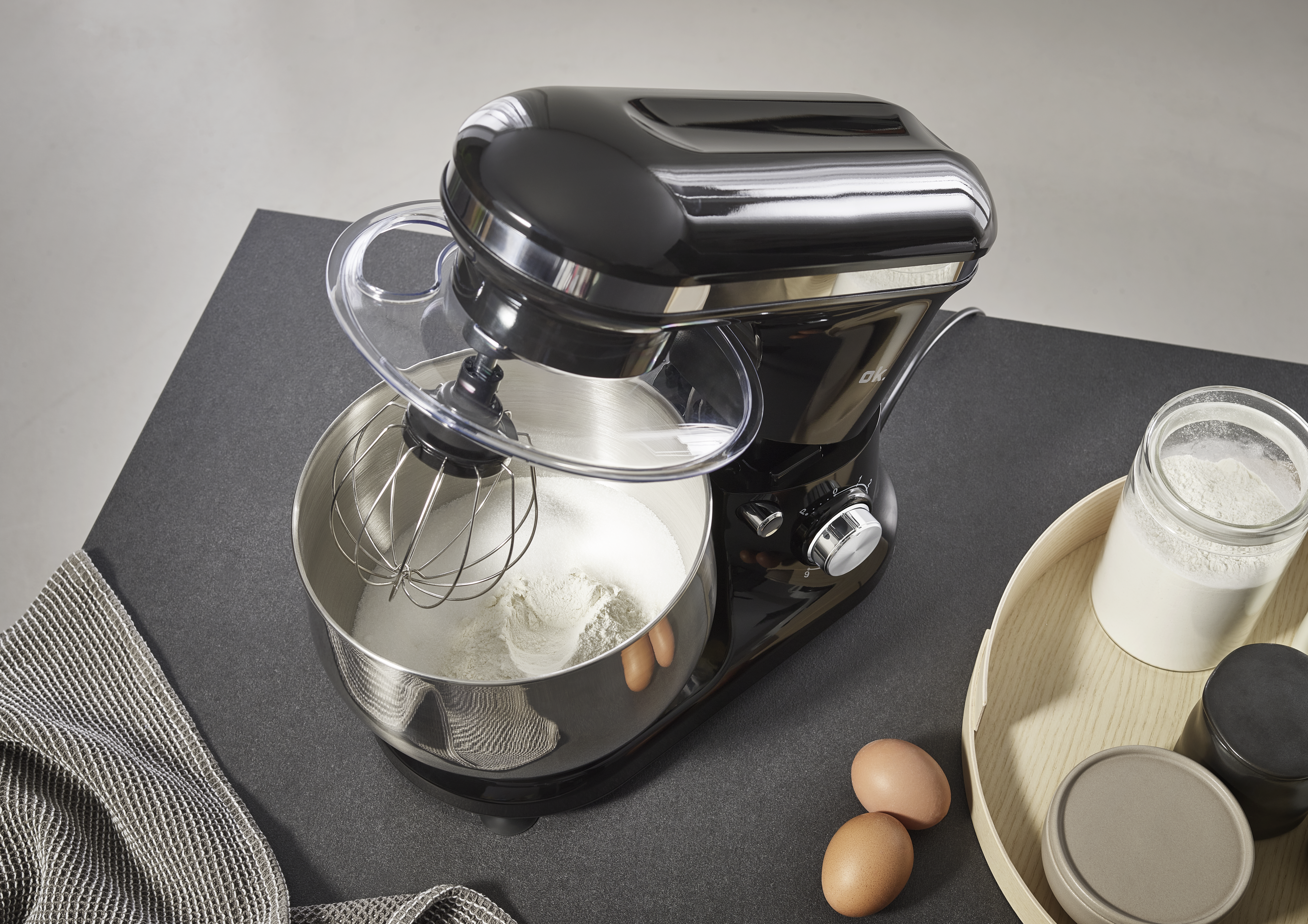 Ein schwarzer Küchenmixer mit Zutaten. Mehl, Eier und ein Glas Milch stehen auf dem Tisch. Der Mixer wird benutzt.
