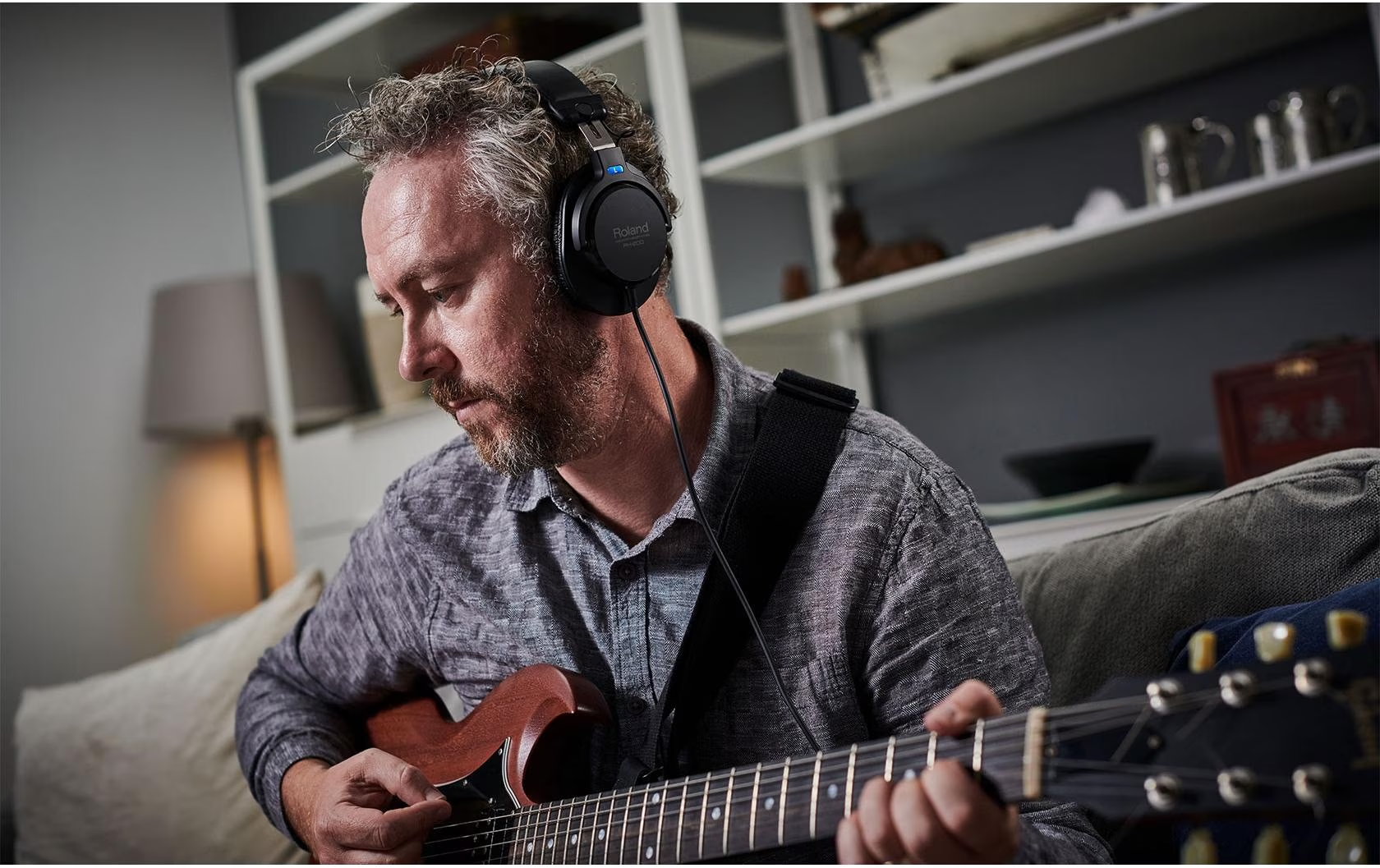 Ein Mann mit Kopfhörern spielt E-Gitarre auf einem Sofa. Im Hintergrund ein Bücherregal.