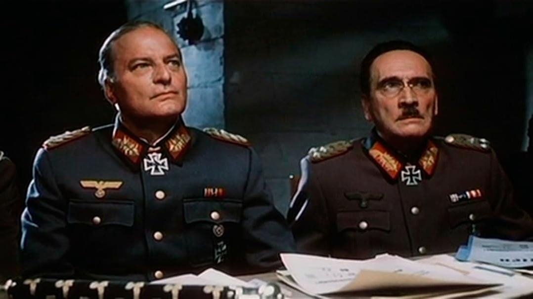 Zwei Männer in Militäruniformen sitzen an einem Tisch. Einer blickt nach oben, der andere trägt eine Brille und einen Schnurrbart.