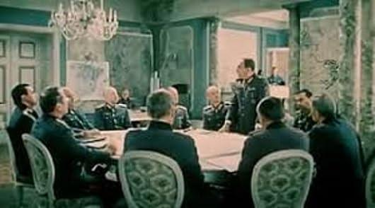 Männer in Uniformen sitzen um einen Tisch. Ein Mann steht und spricht. Eine Leuchte hängt darüber. Der Raum hat eine verzierte Dekoration.