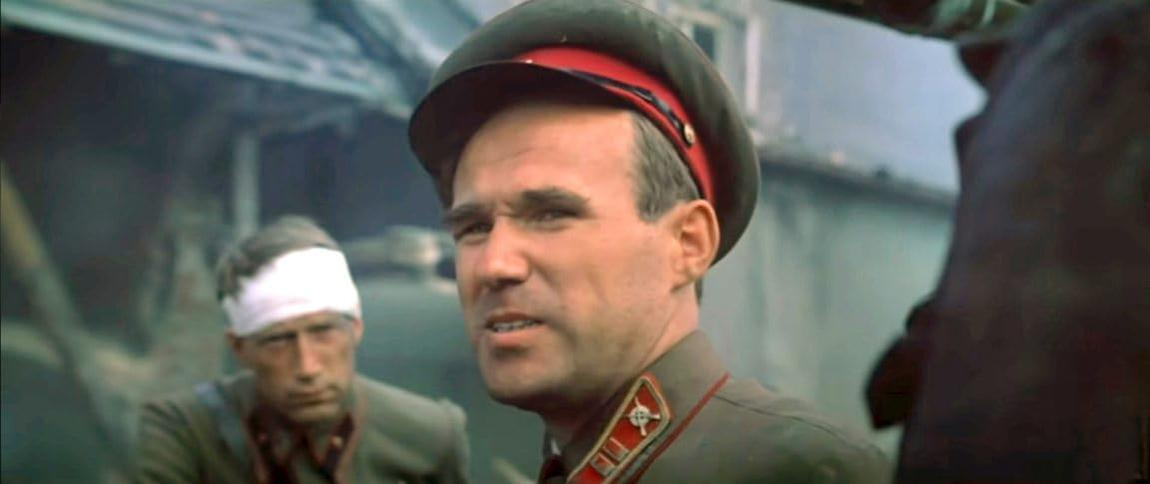 Ein Mann in Uniform und Hut spricht. Ein anderer Mann mit Kopfverband ist im Hintergrund.
