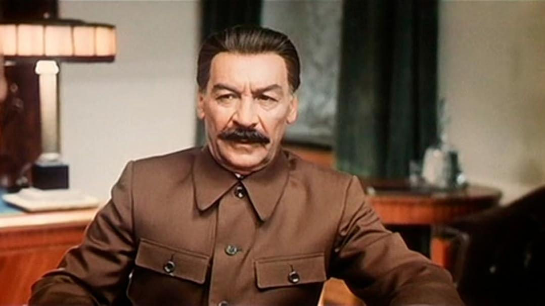 Ein Mann in brauner Uniform mit Schnurrbart, möglicherweise Stalin, blickt in die Kamera. Der Hintergrund hat einen Tisch und eine Lampe.