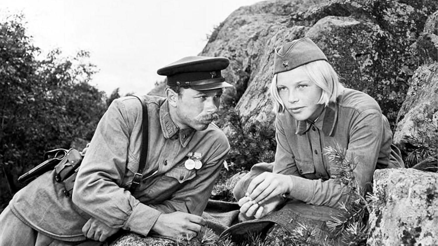 Ein Mann und eine Frau in Militäruniformen sitzen zusammen im Freien. Der Mann hat einen Schnurrbart, eine Mütze und Medaillen. Die Frau hat kurze Haare.