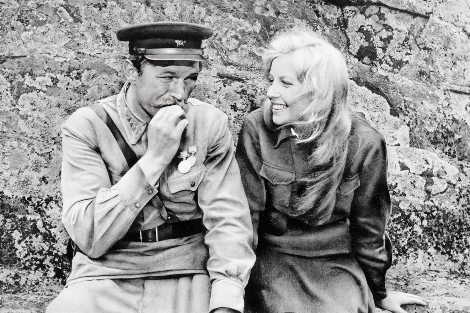 Ein Mann in Uniform und eine Frau mit blonden Haaren sitzen nebeneinander und lächeln vor einem Felsenhintergrund.
