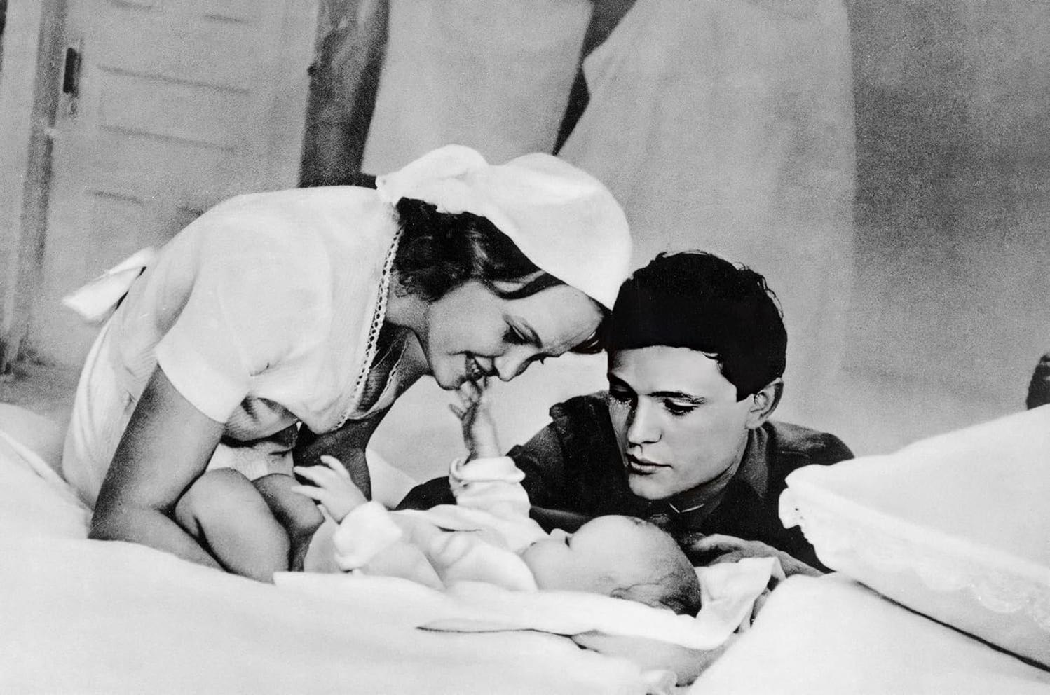Eine Frau und ein Mann betrachten ein Baby, das auf einem Bett liegt. Die Frau trägt eine Mütze und der Mann schaut zu.