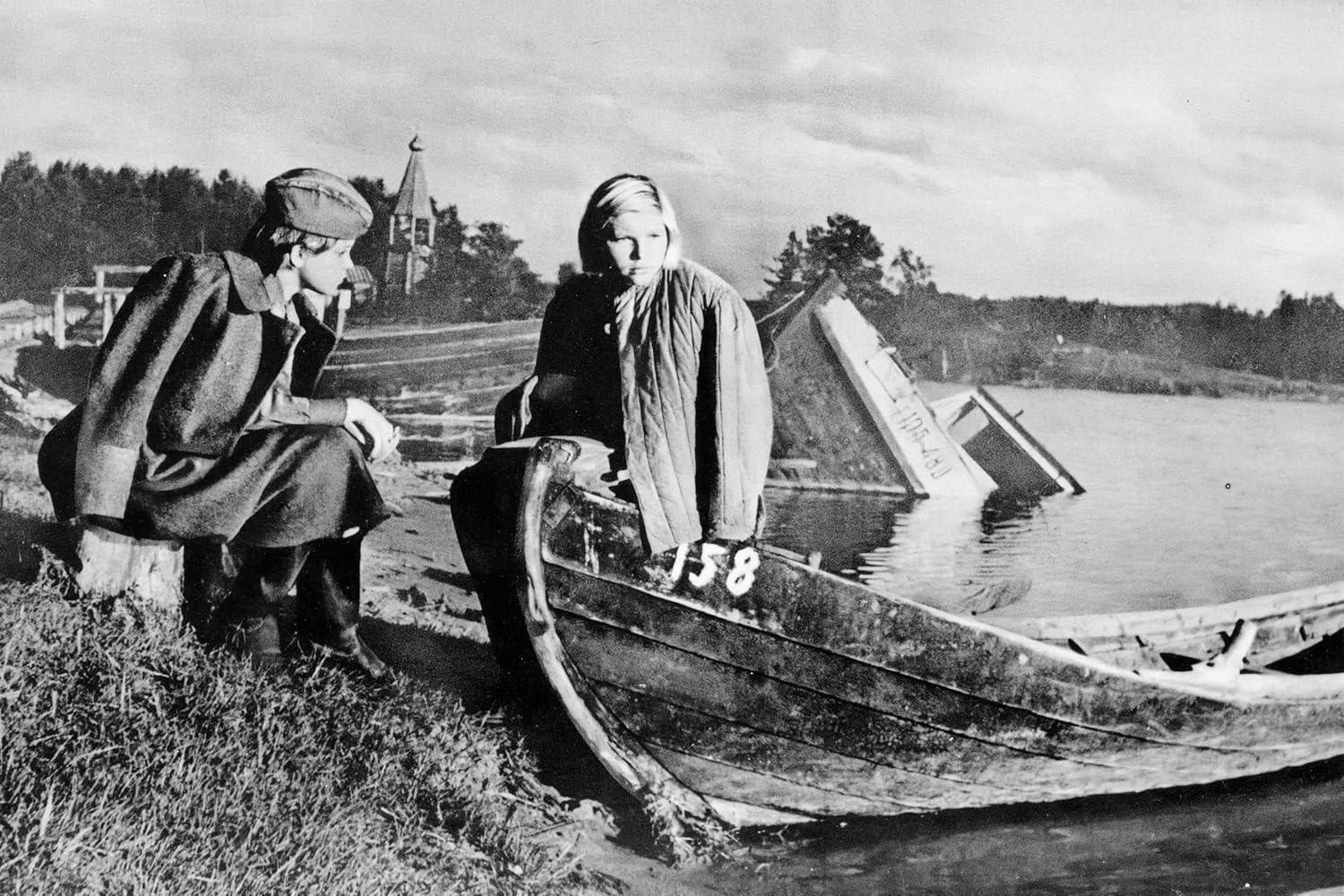 Zwei Personen sitzen in der Nähe eines Bootes am Wasser, im Hintergrund eine Kirche und Bäume. Eine Person schaut hinaus, die andere sitzt im Boot.