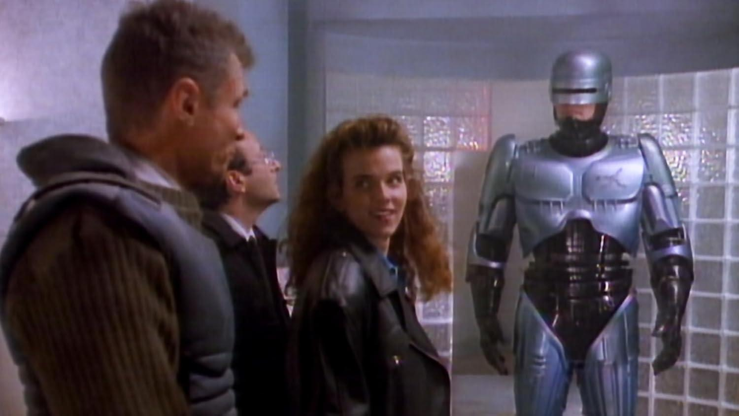 RoboCop steht inmitten einer Gruppe von Menschen. Eine Frau lächelt. Zwei Männer in Anzügen blicken nach oben. Hinter ihnen ist eine Glasblockwand.