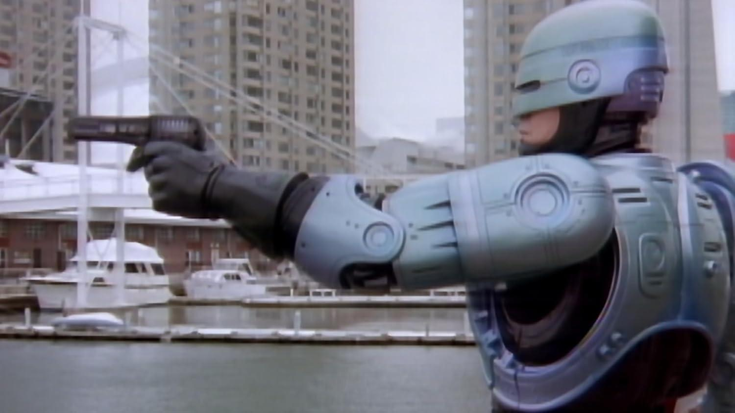 RoboCop zielt mit einer Waffe. Er befindet sich in einer Stadt, in der Nähe von Wasser mit Booten. Im Hintergrund sind Gebäude und der Himmel ist bedeckt.