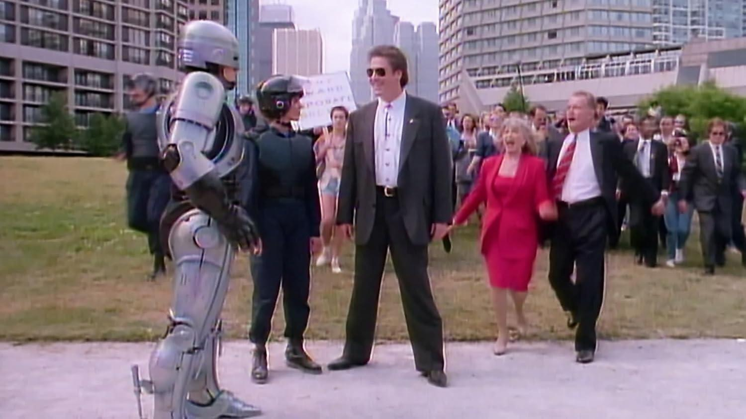 RoboCop steht einer Gruppe von Menschen gegenüber, darunter Anzüge und eine Frau in Rot, in einem Park. Hintergrund Gebäude und Himmel vorhanden.