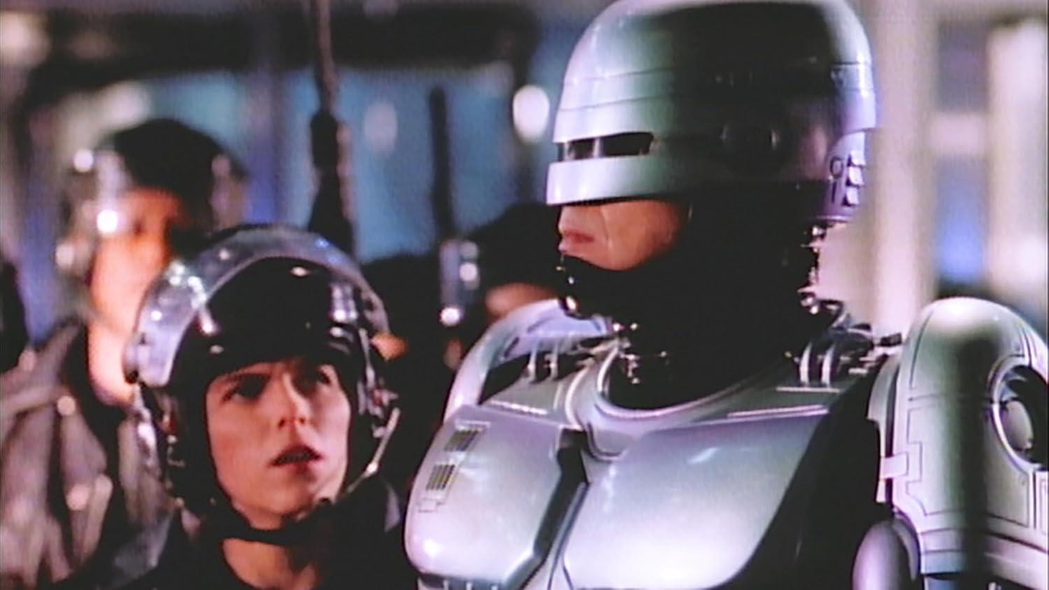 RoboCop steht neben einem Polizisten. Beide tragen Helme. Im Hintergrund sind andere Beamte und eine verschwommene Innenszene zu sehen.