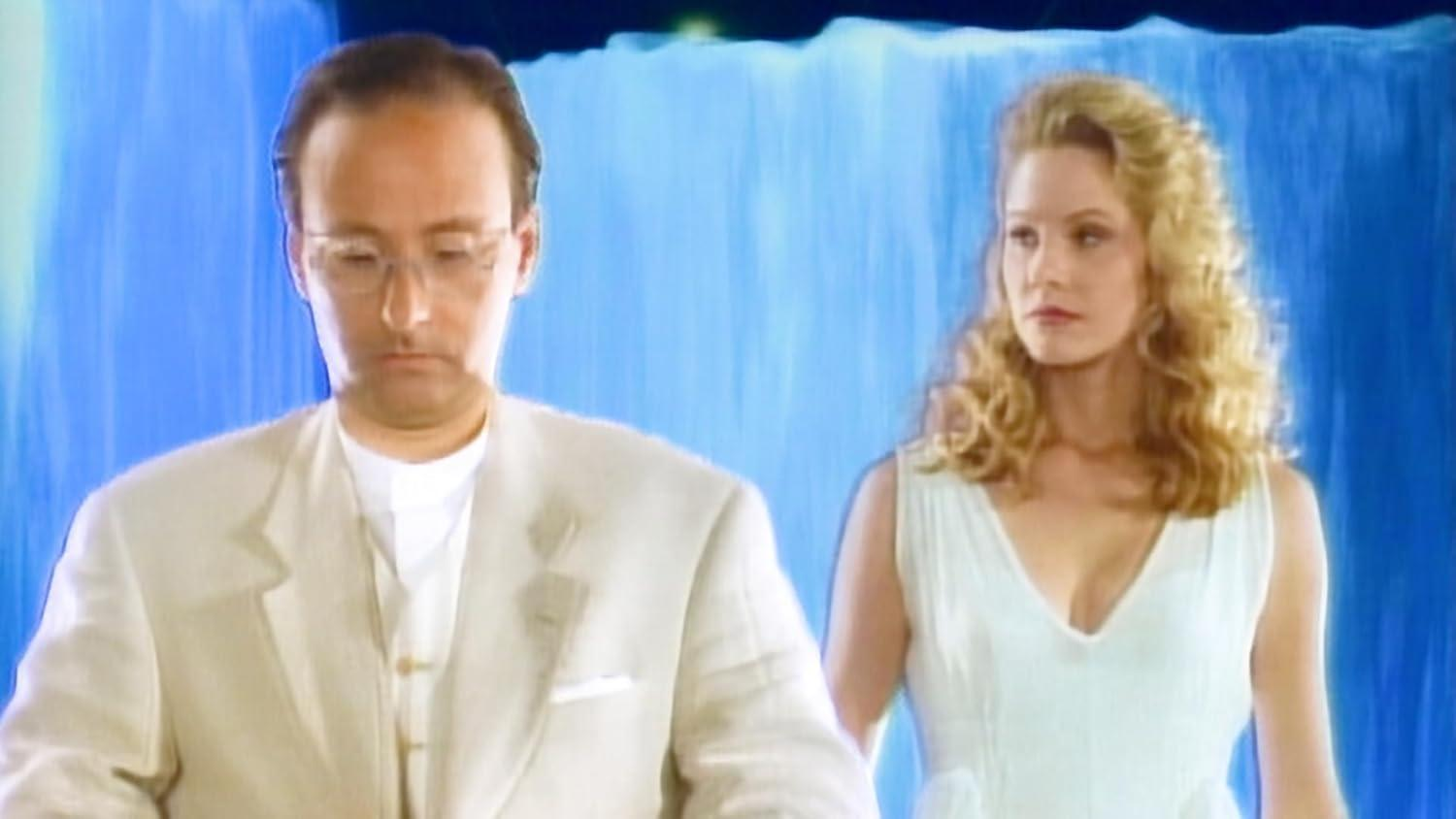 Ein Mann in hellem Anzug und eine Frau in weißem Kleid stehen vor blauem Hintergrund. Der Mann trägt eine Brille.